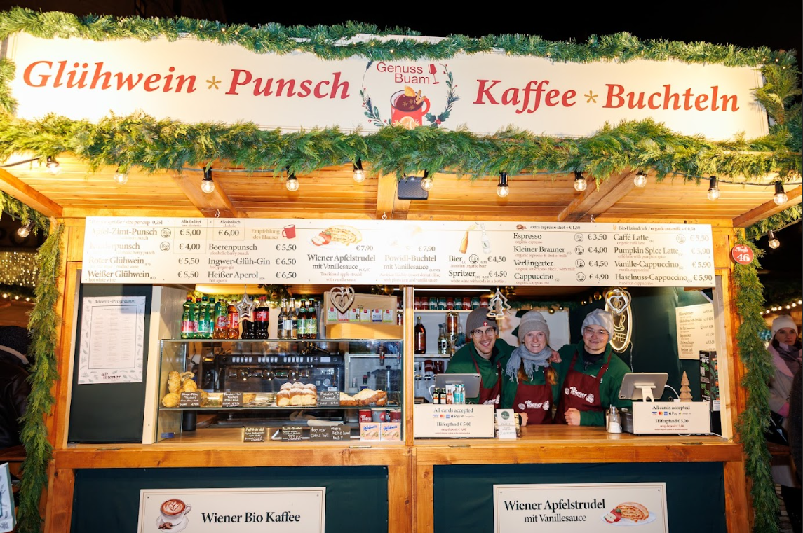 Festive Christmas market stall adorned with greenery serves Glühwein, Punsch, coffee, and Buchteln. Three smiling vendors in red aprons stand ready to assist customers.