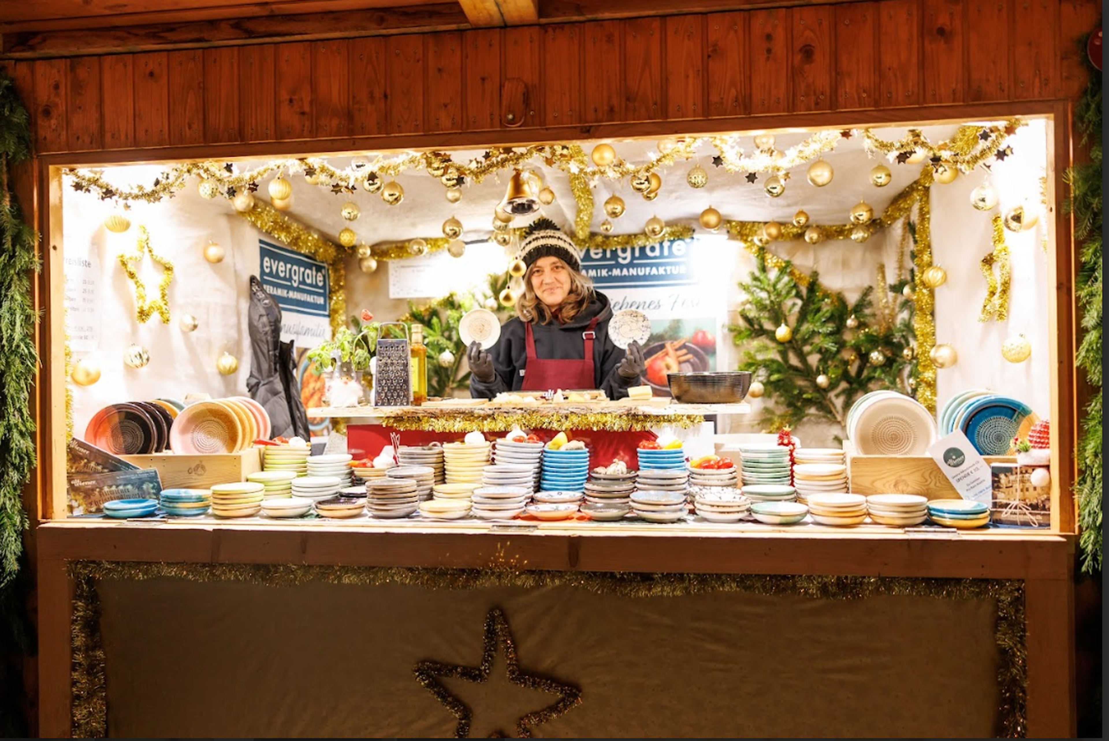 A person smiles at a festive market stall filled with colorful ceramic bowls and plates. The booth is adorned with gold ornaments and garlands.