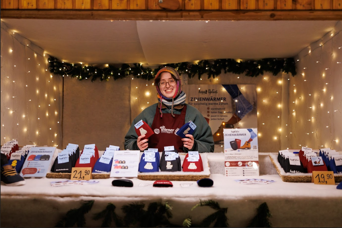 A vendor at a festive market stall adorned with warm lights displays colorful packages of items on a counter. Prices are visible, creating a cozy, inviting atmosphere.