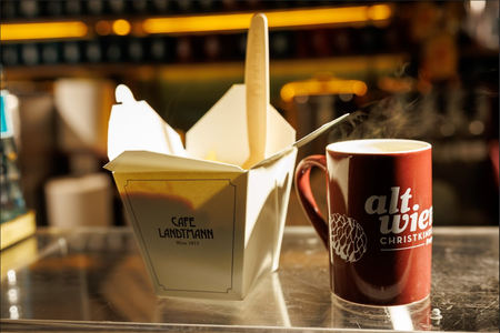 A steaming mug with "alt.wien Christkindl" text sits beside a takeout box labeled "Cafe Landtmann, Wien 1873" on a reflective surface, in warm light.