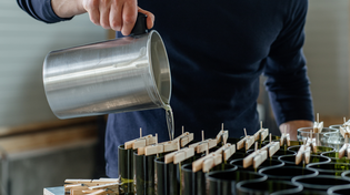 Person pouring melted wax into green candle molds, with wooden wicks secured by clothespins, on a worktable.