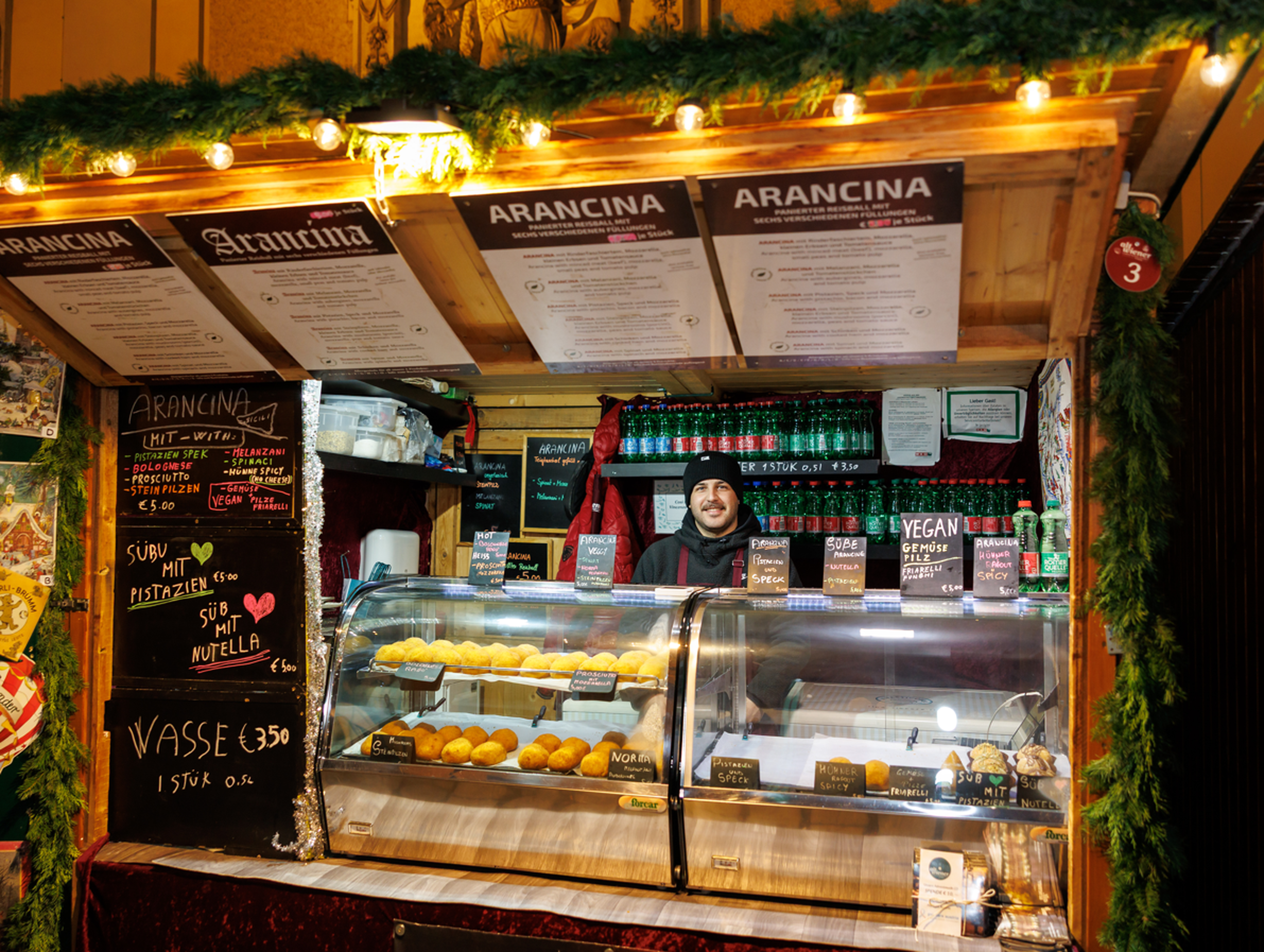 A wooden food stall at a Christmas market decorated with lights and greenery, where a smiling vendor stands behind a glass display filled with arancini. Menus and chalkboard signs showing flavors and prices are visible around the counter.