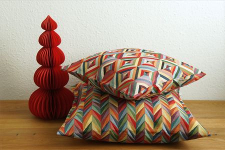 Two colorful, geometric-patterned pillows stacked on a wooden table next to a red, cone-shaped paper decoration.