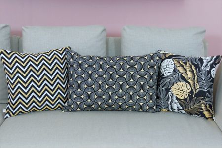 Three decorative pillows with yellow, black, and white patterns are arranged on a beige sofa against a pink wall.