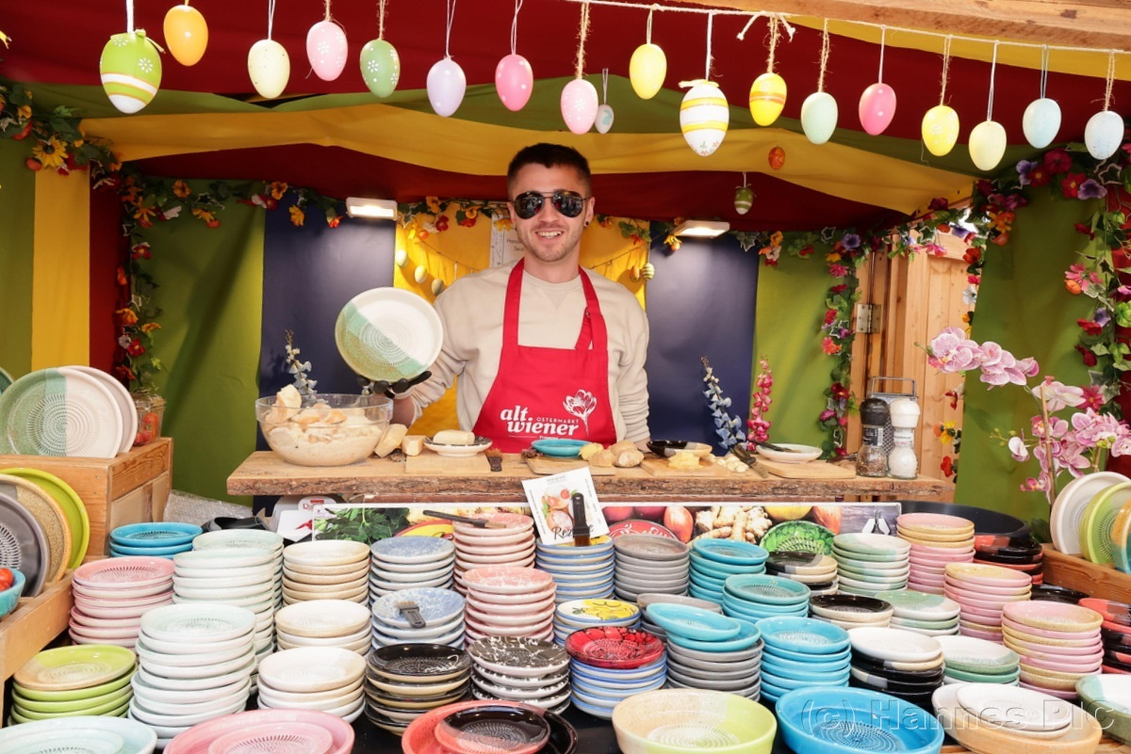 A vendor in sunglasses and a red apron selling colorful ceramic plates at a festive market stall decorated with hanging Easter eggs.