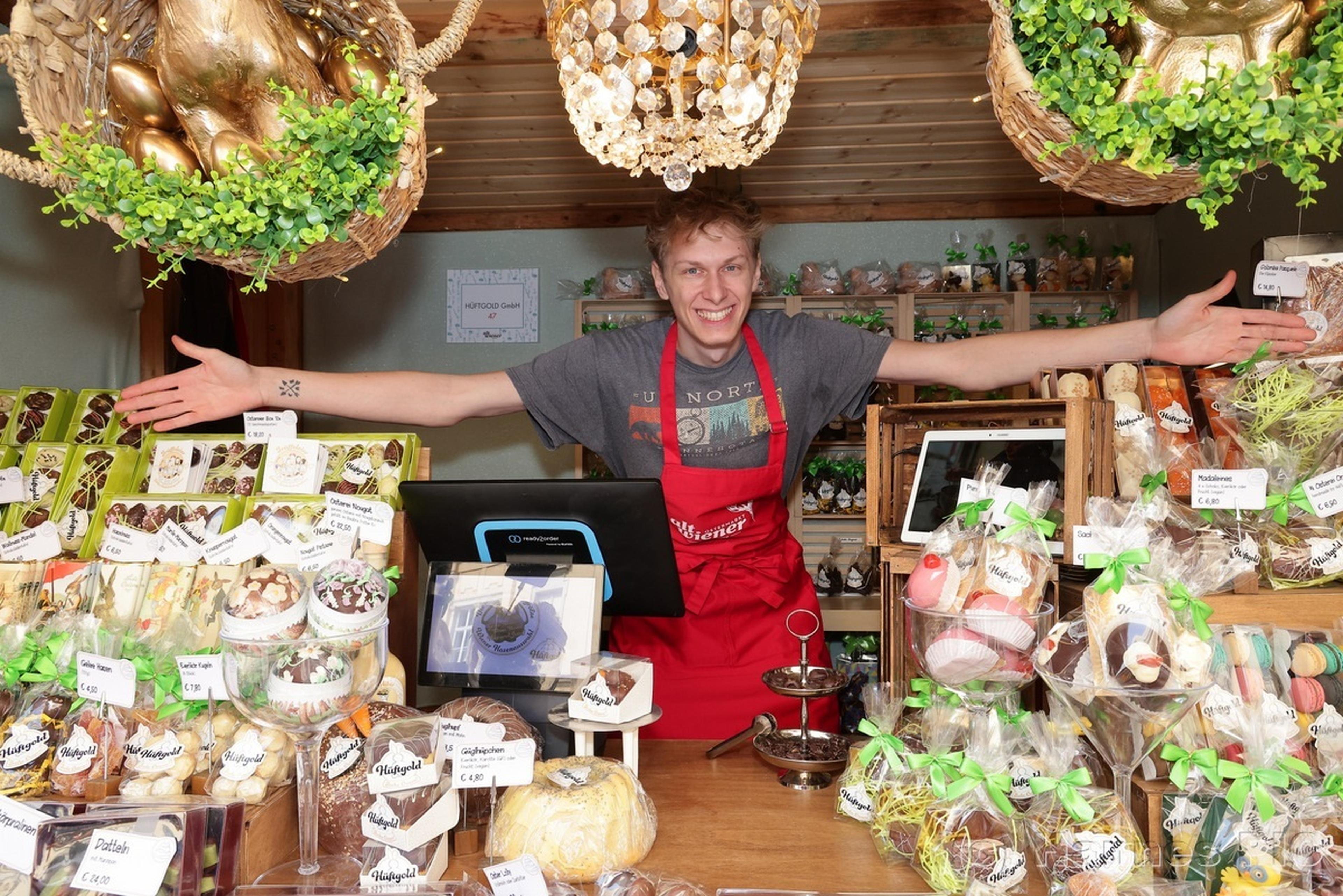 A smiling vendor in a red apron stands with arms outstretched behind a display of assorted baked goods in a cozy, decorated shop.