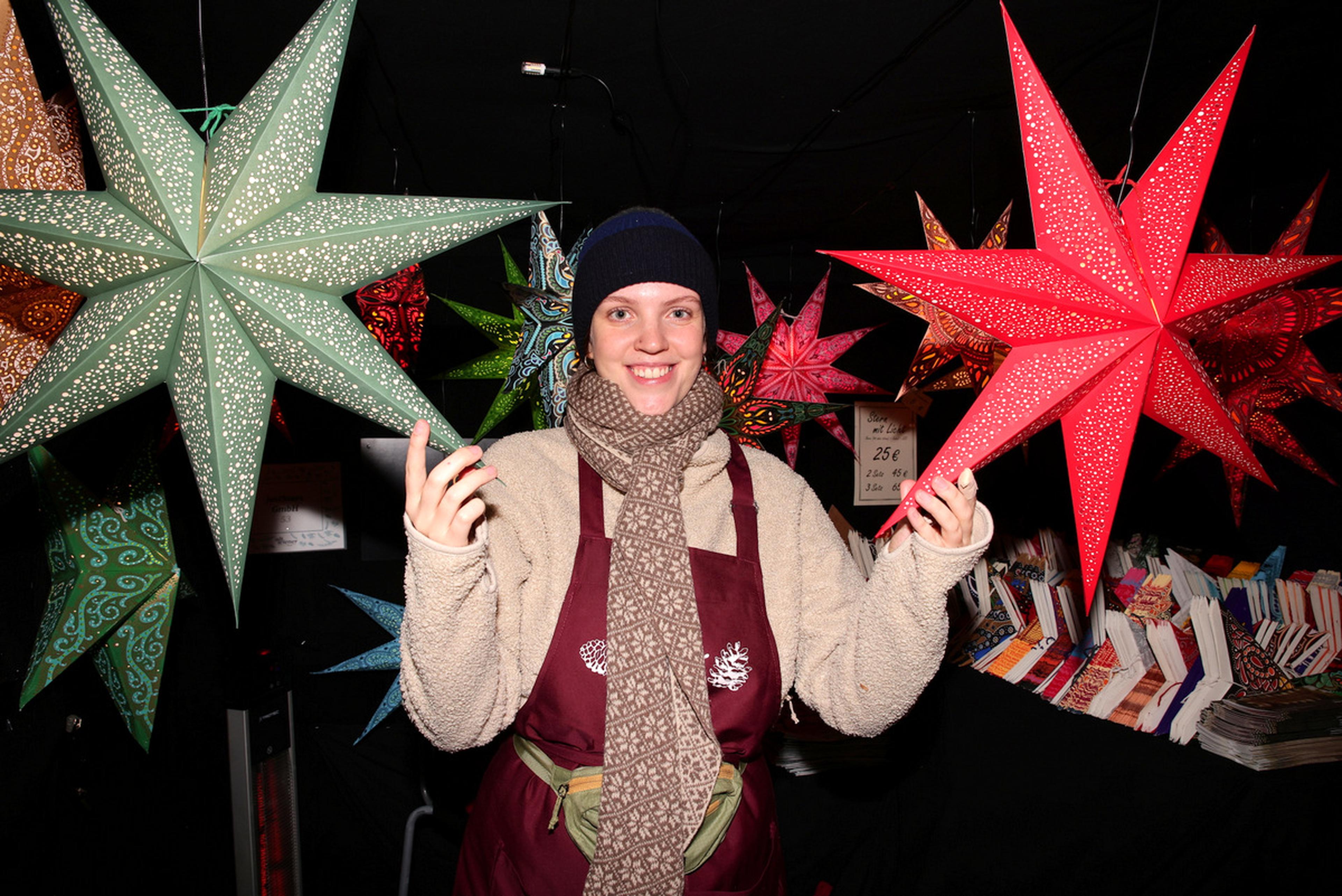 A person in a scarf and hat smiles, holding colorful star lanterns at a market stall with more stars and books in the background.