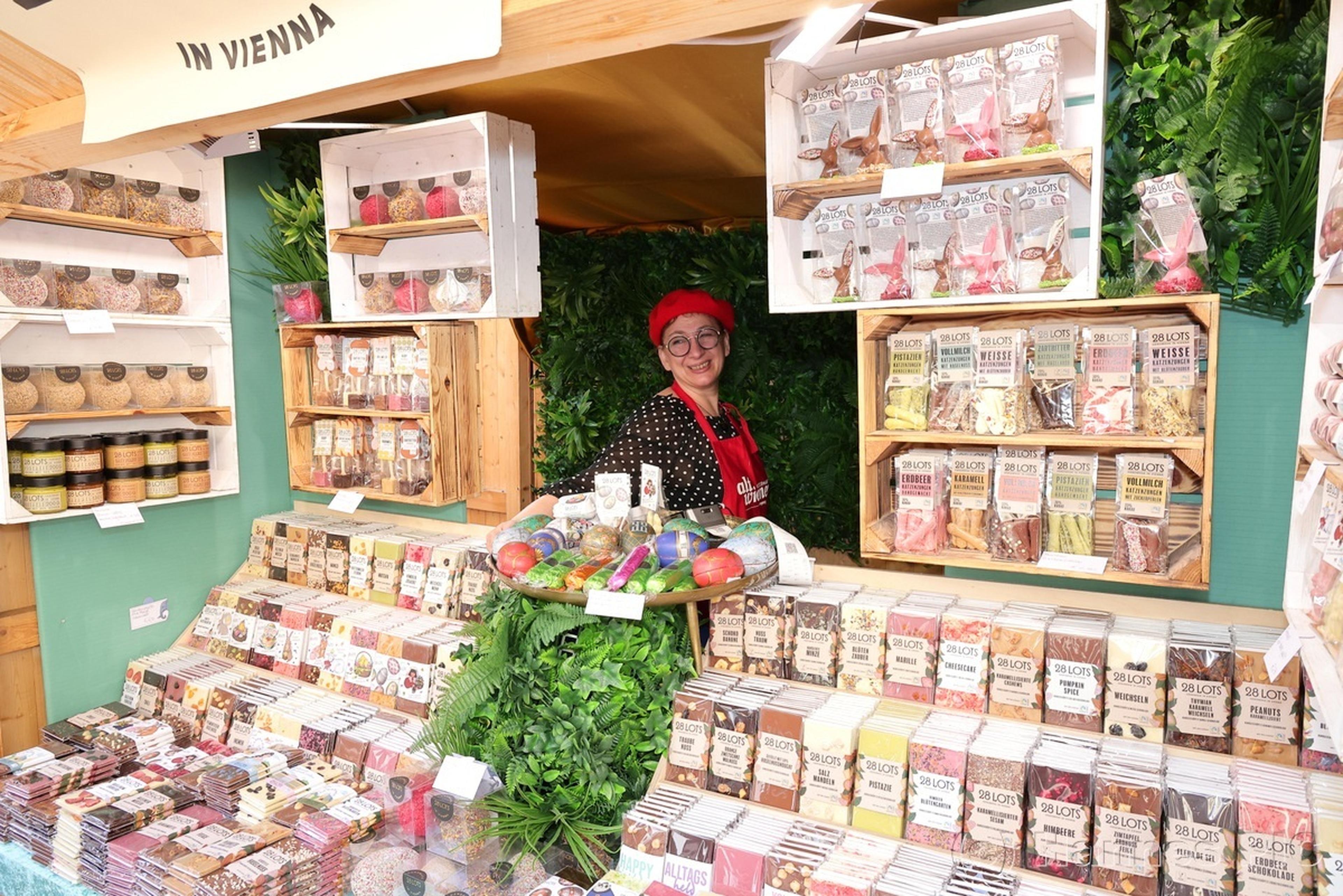 A vendor in a red beret stands at a colorful stall selling assorted chocolates, surrounded by greenery and shelves of treats.