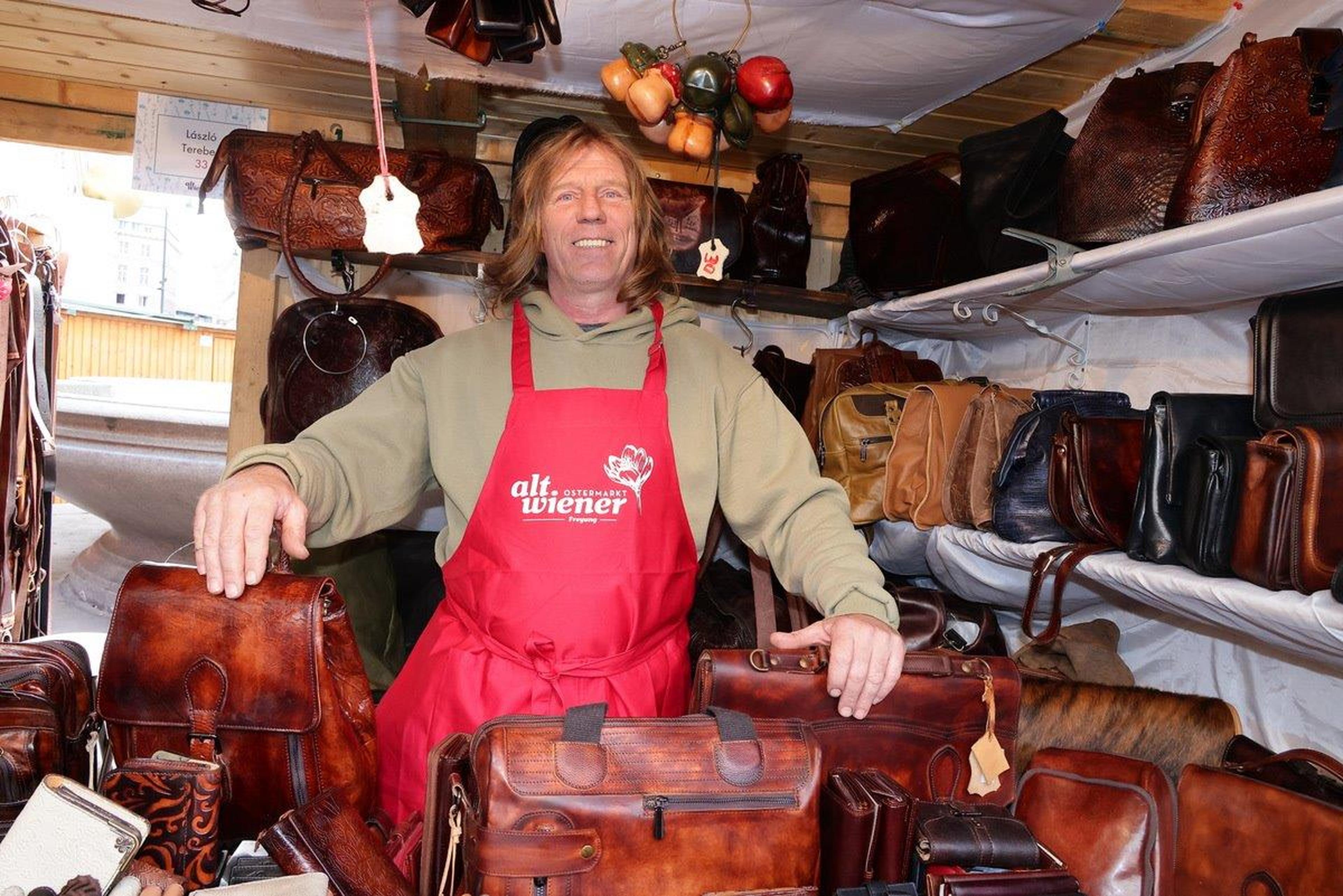 A vendor in a red apron stands smiling in a stall filled with various leather bags.