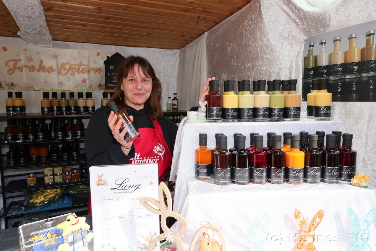 A woman in an apron holds a bottle, showcasing a display of colorful bottles at a market stall with "Frohe Ostern" sign in the background.