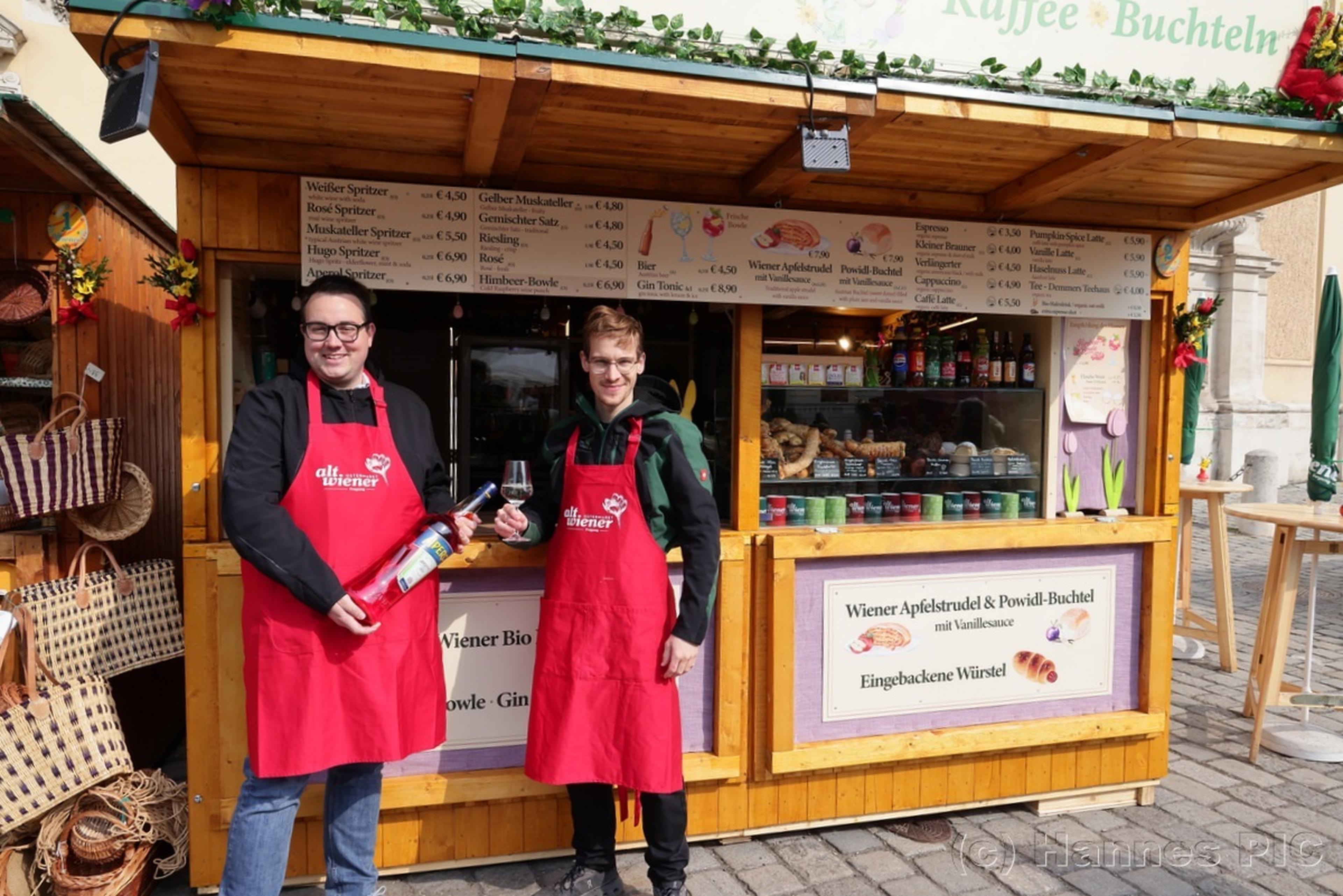 Two smiling vendors in red aprons stand in front of a wooden market stall, holding wine bottles and glasses, with shelves of products behind them.