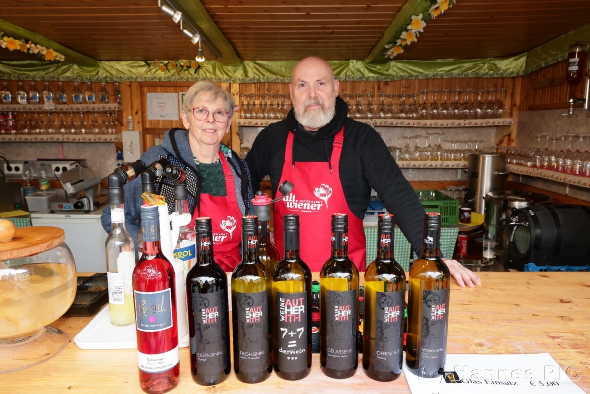 A man and woman stand behind a counter with various wine bottles displayed in front of them, inside a cozy wooden wine shop.