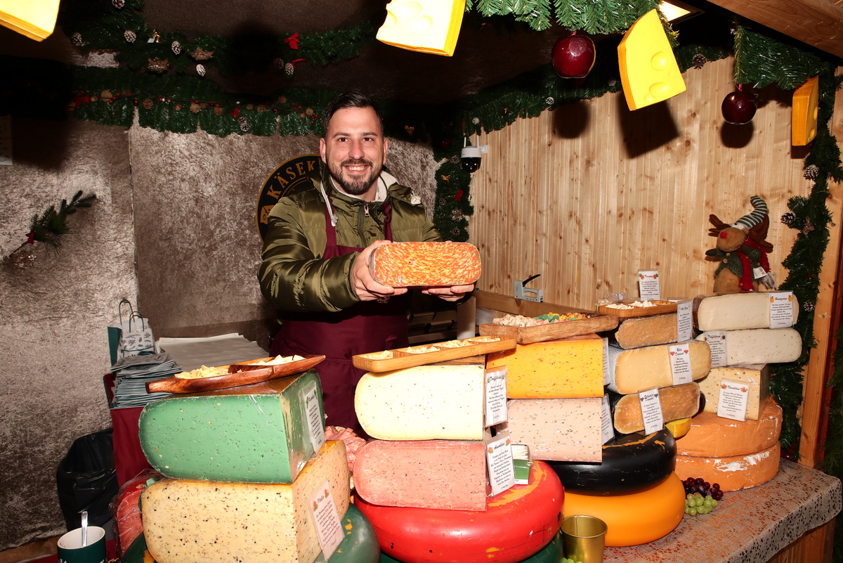 A vendor in a festive market stall holds a cheese wheel, surrounded by various colorful cheeses and decorations.