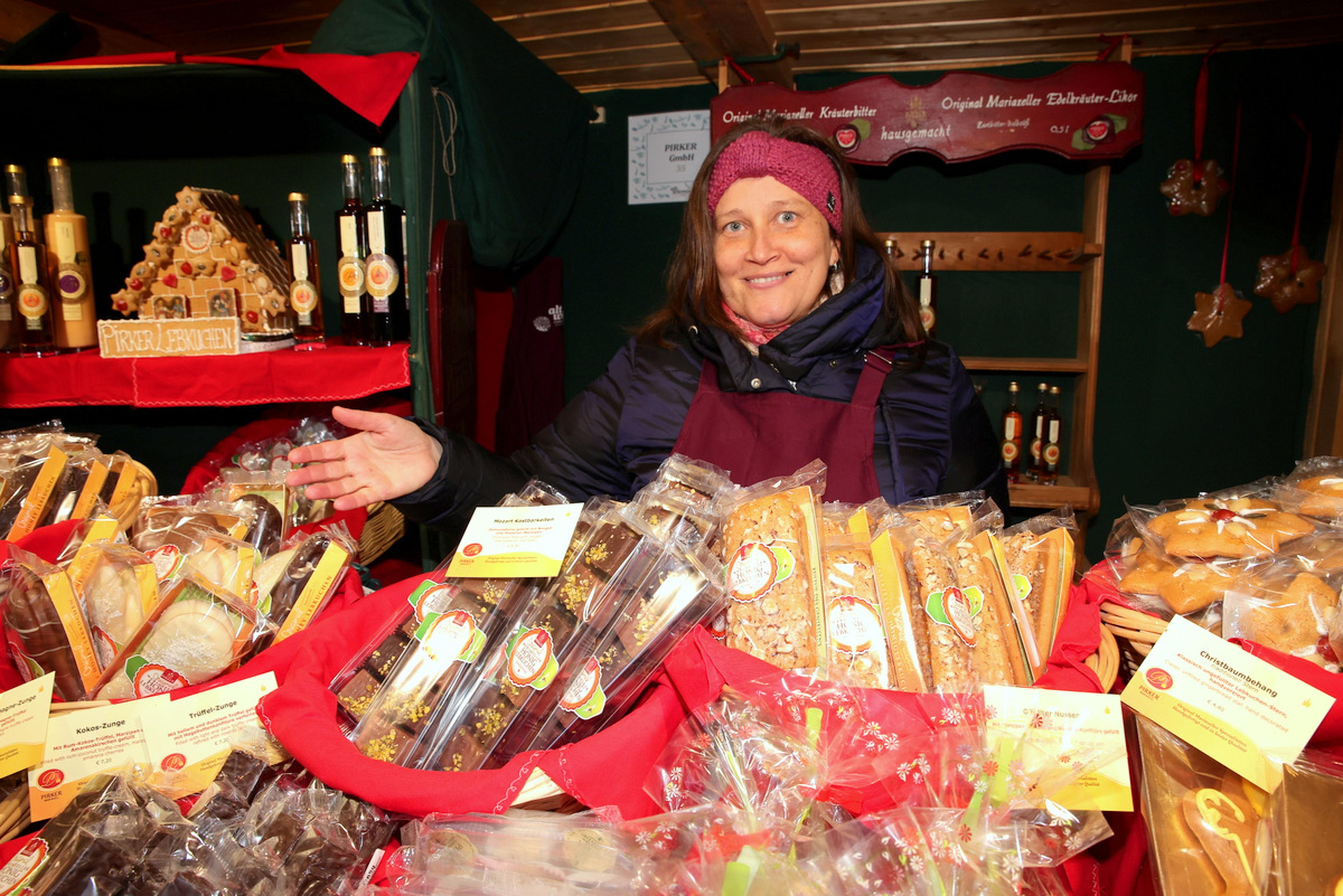 A vendor in a cozy hat and apron smiles while displaying a variety of packaged baked goods at a festive market stall.