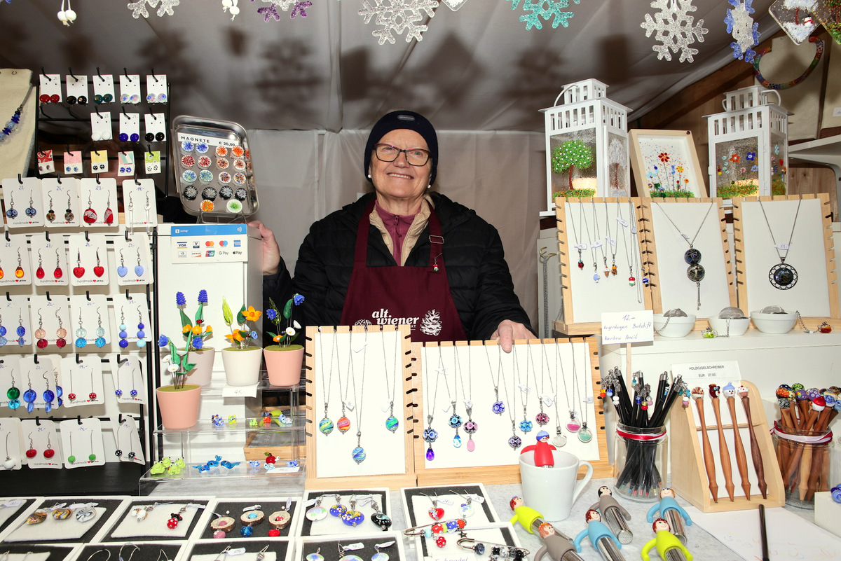 A person stands in a booth displaying handmade jewelry, including necklaces and earrings, surrounded by decorative snowflakes.