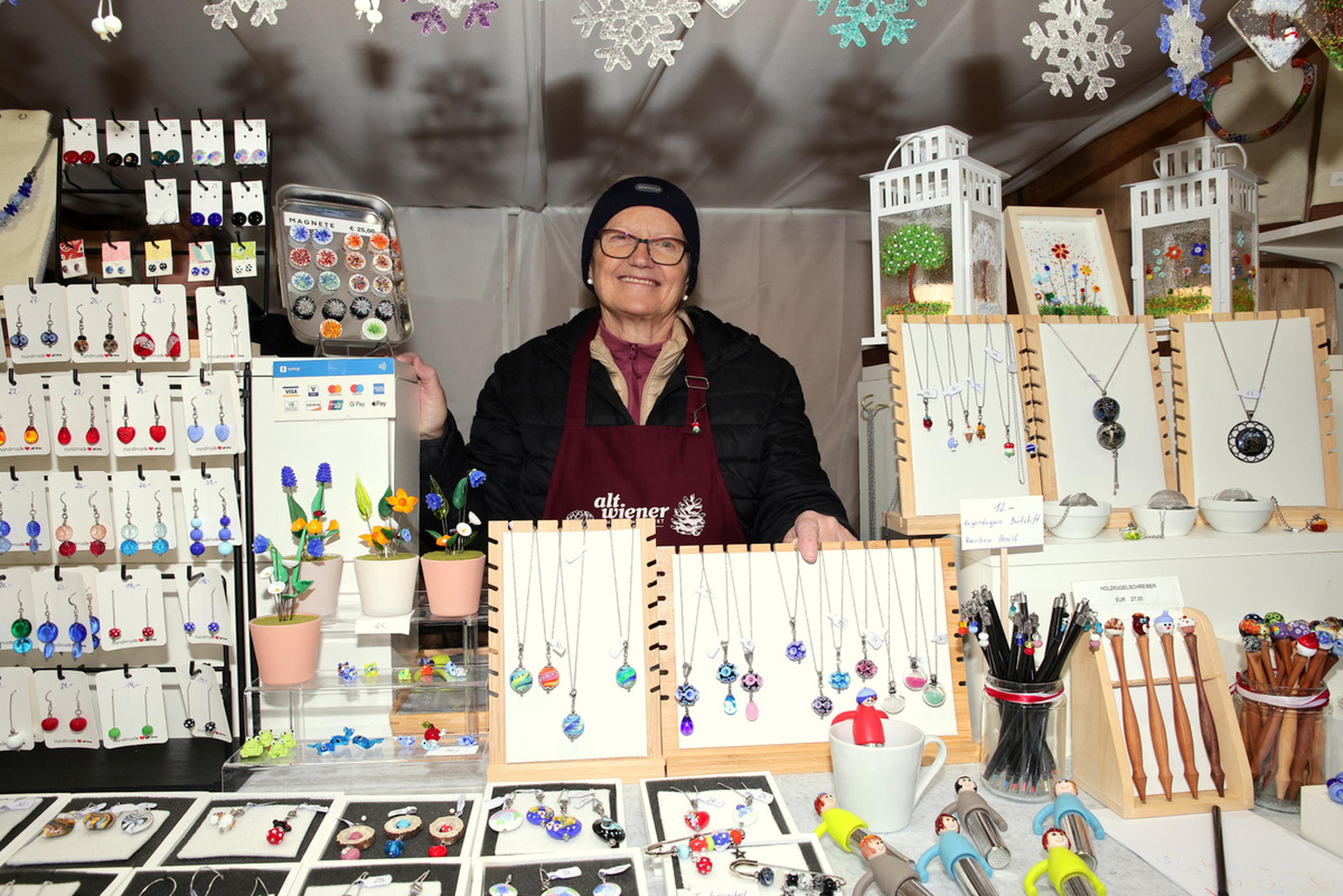 A person stands in a booth displaying handmade jewelry, including necklaces and earrings, surrounded by decorative snowflakes.