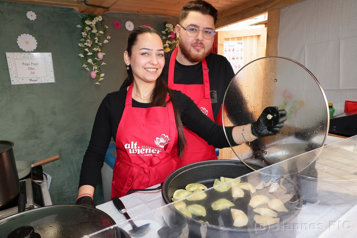 Two people in red aprons cooking dumplings in a pan, one holds the lid. The booth backdrop has a floral decoration.