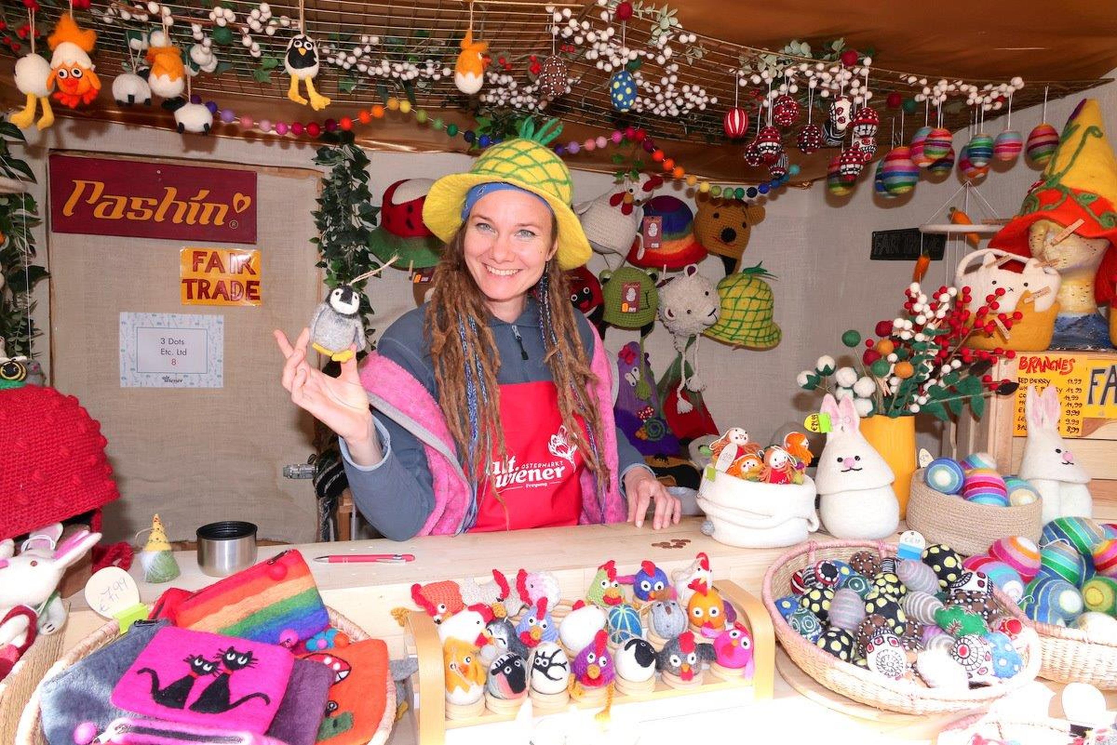 A smiling vendor in a colorful hat sells handmade knitted toys and decorations at a festive market stall.