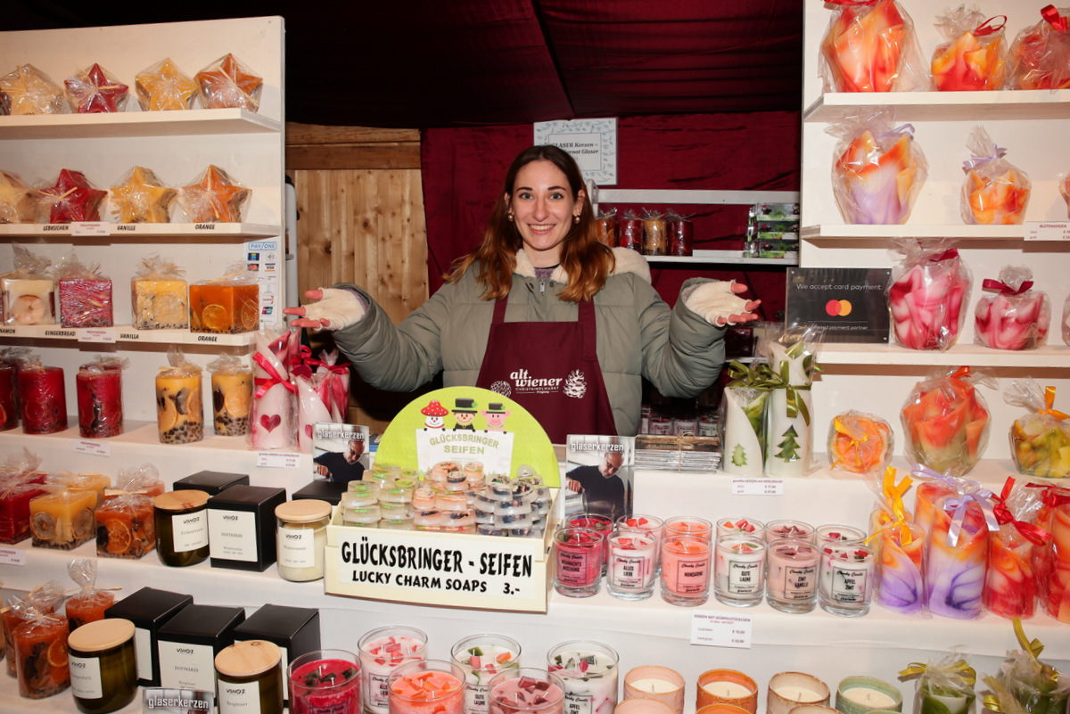 A woman in an apron stands smiling behind a display of colorful candles and soaps in a wooden booth.