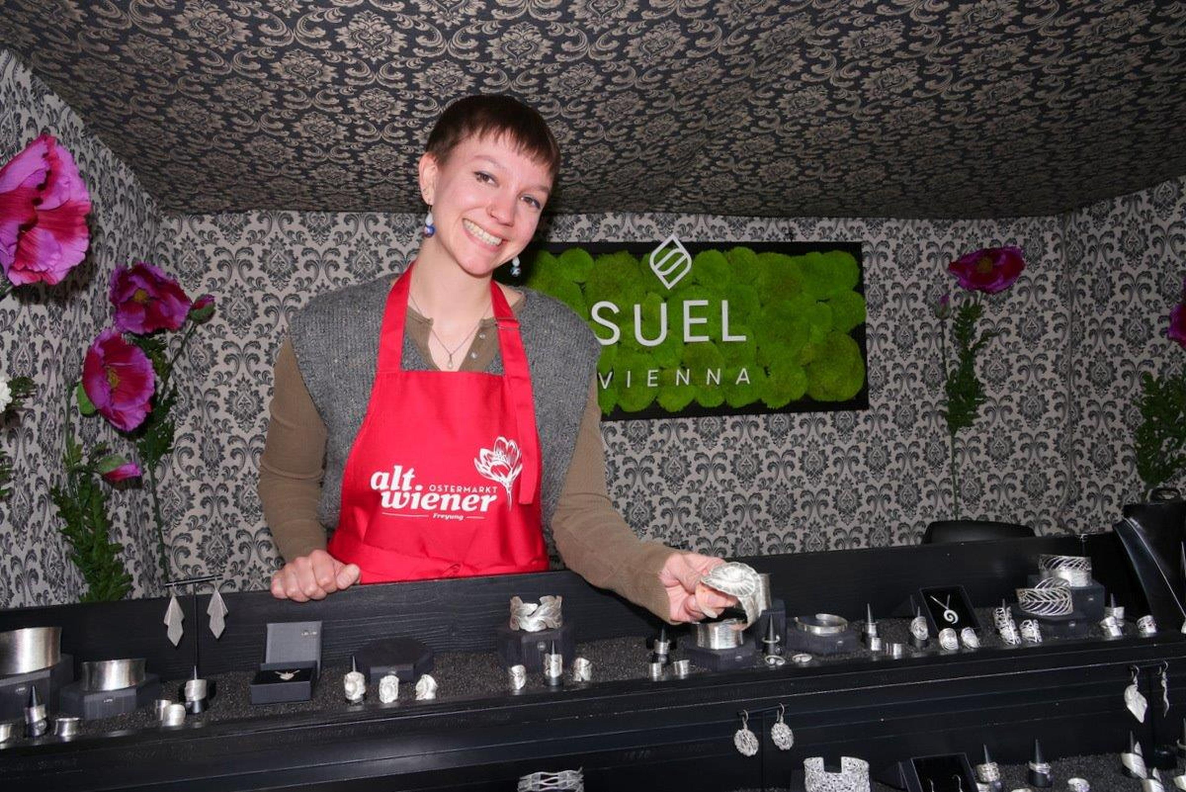 Person in a red apron stands behind a jewelry display, holding a silver ring. Floral and patterned backdrop with "Suel Vienna" signage.