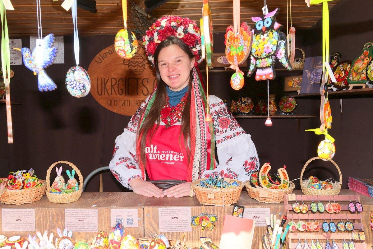 A woman in traditional Ukrainian attire stands behind a crafts stall, displaying colorful handmade decorations and folk art items.