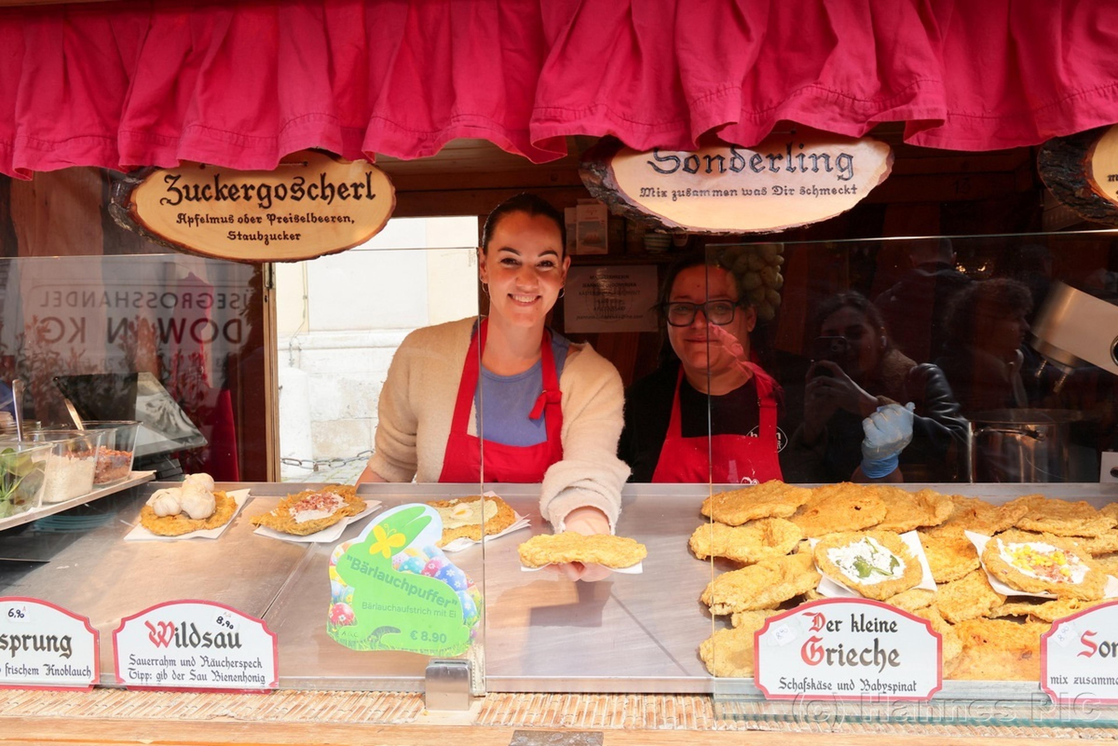 Two smiling vendors at a food stall display various fried dishes, with signs in German describing the delicacies.