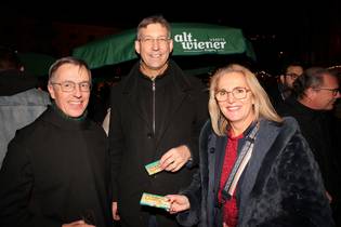 Three people smiling and holding cards at an outdoor event, with a green umbrella and festive lights in the background.