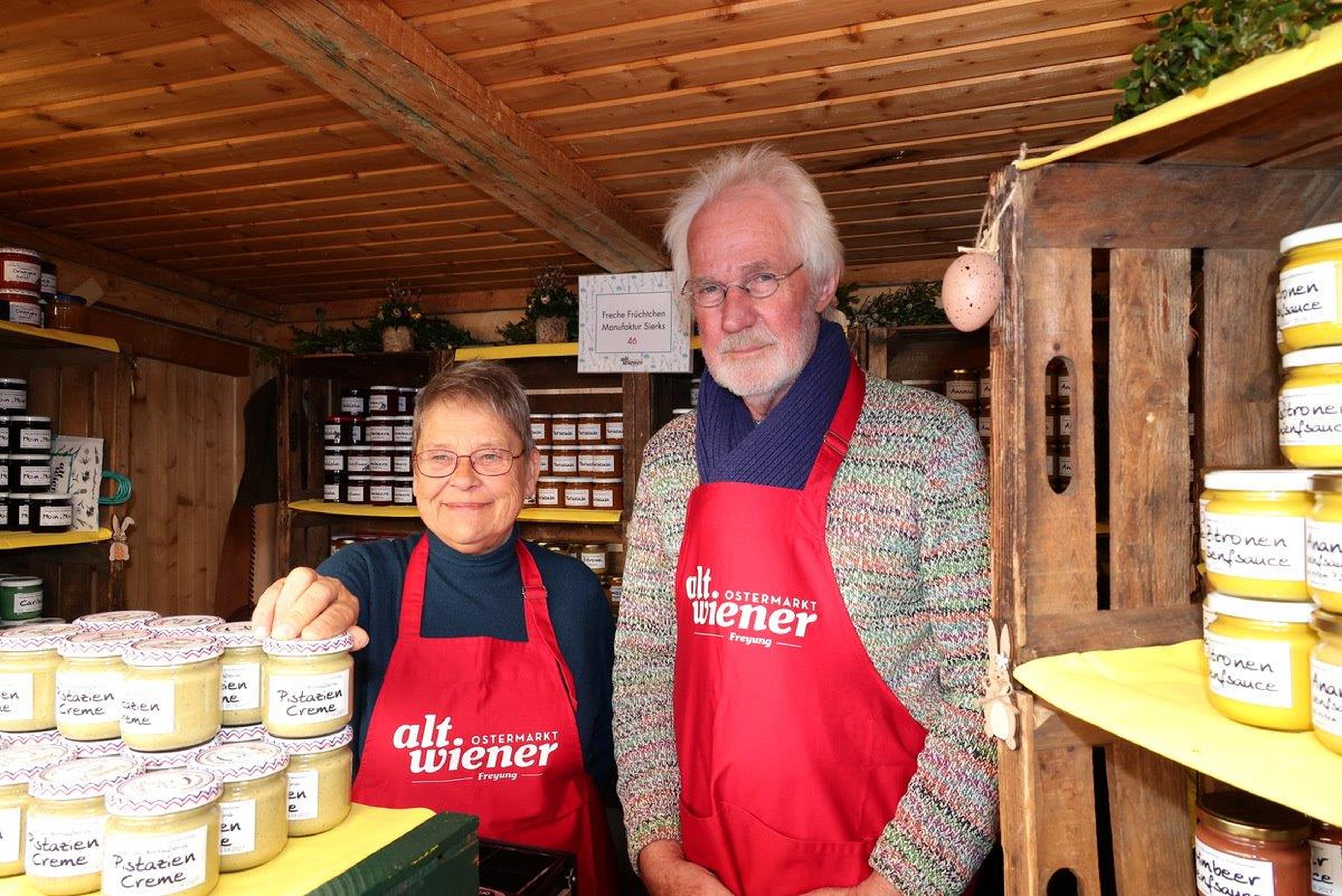 Two people in red aprons stand in a wooden booth with shelves of jars, smiling and presenting their products.