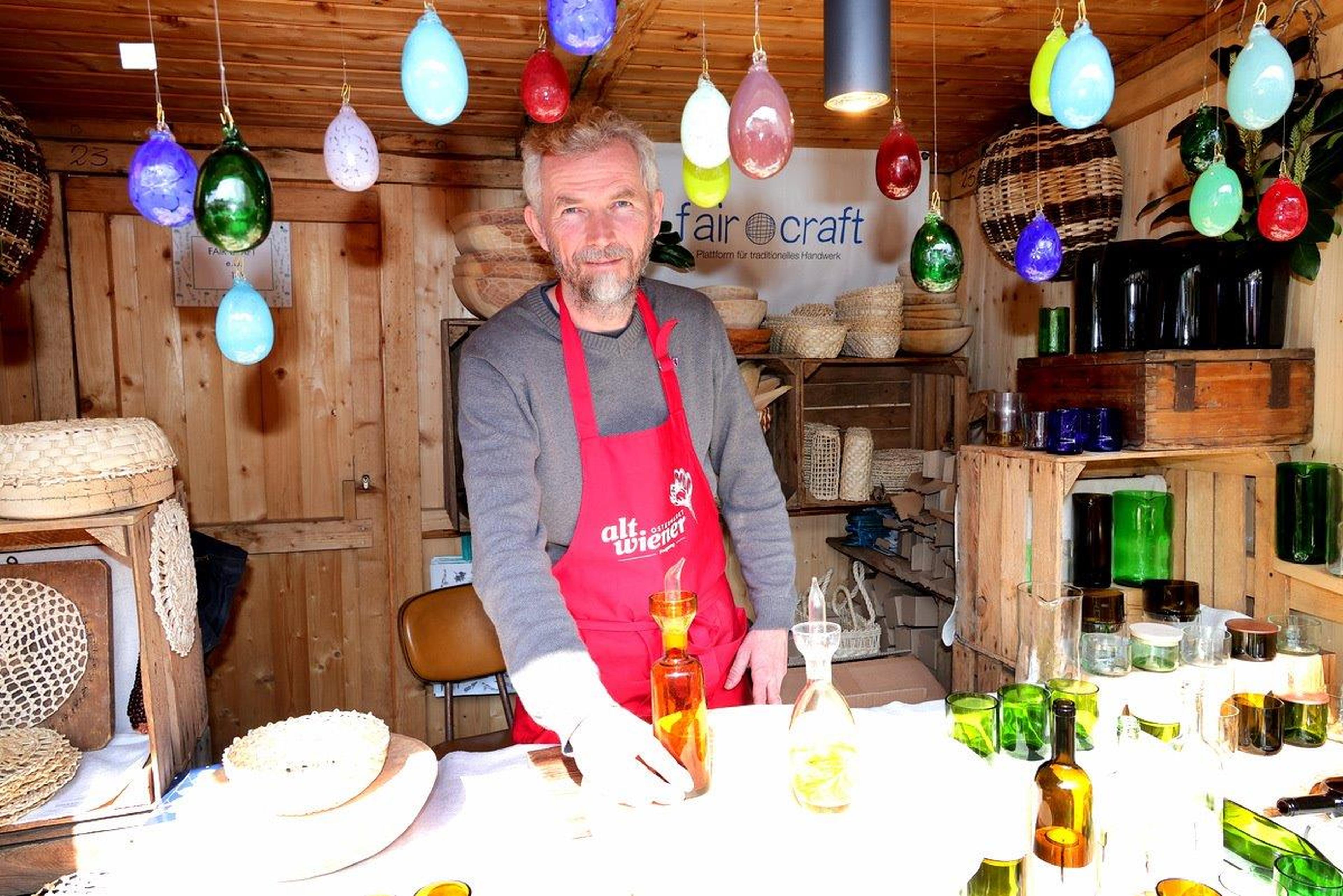 Man in a red apron at a wooden stall with colorful hanging lights and glassware, showcasing handmade crafts.