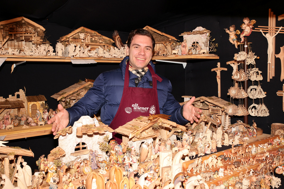 Person wearing a maroon apron stands smiling behind a display of detailed nativity scenes and wooden figures at a market stall.