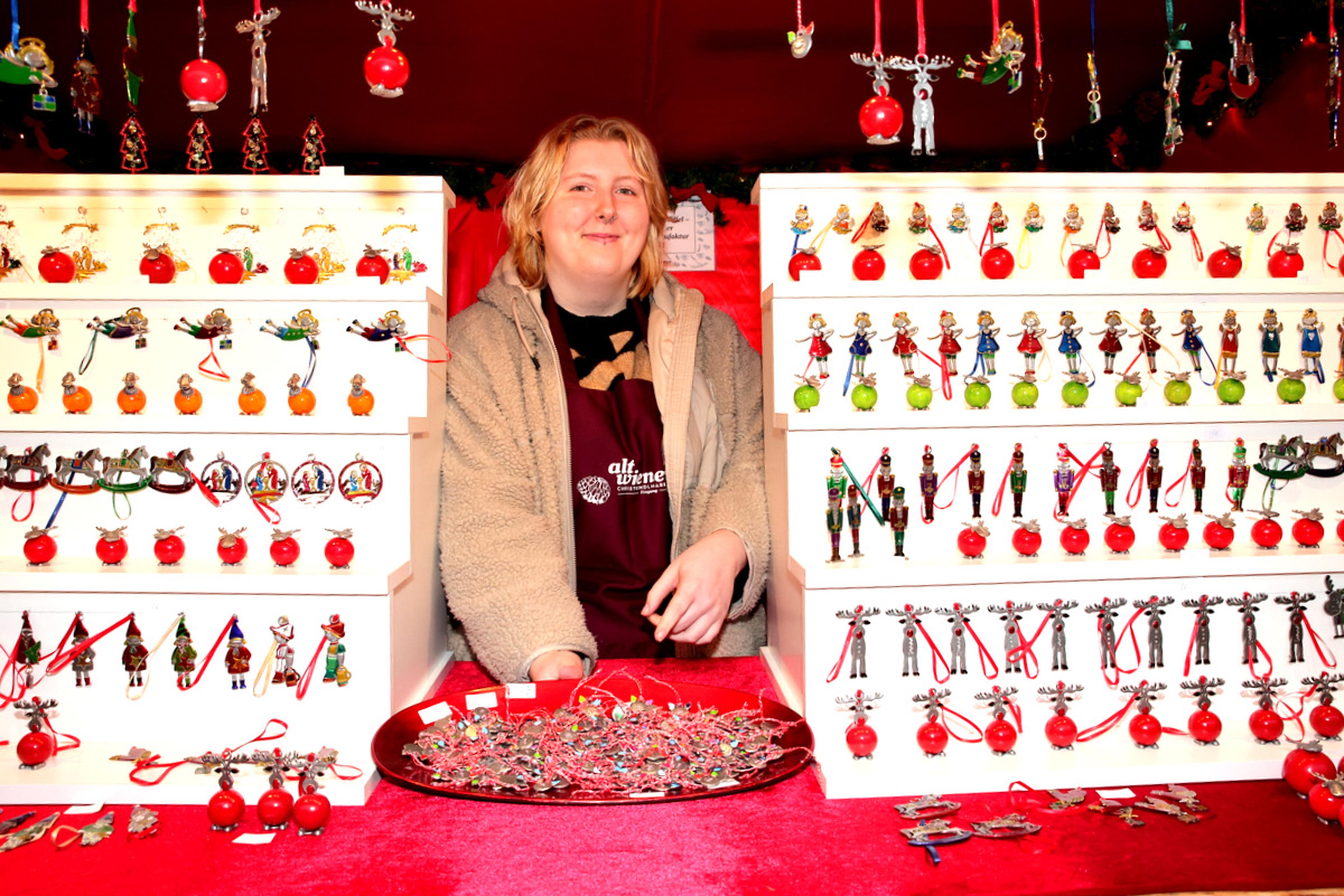 Person at a festive market stall displaying various colorful Christmas ornaments and decorations on white shelves, with a red backdrop.