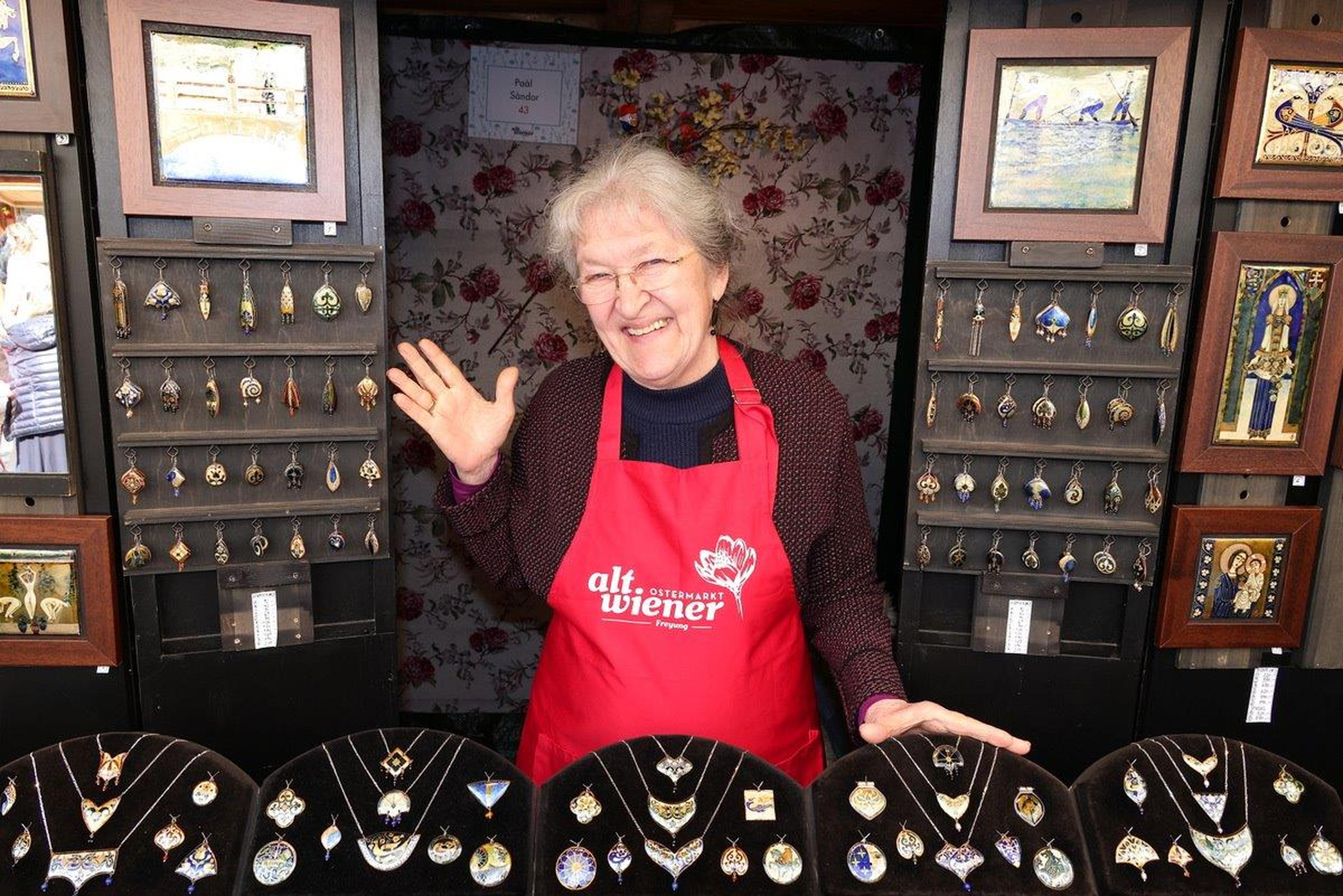 Smiling vendor wearing a red apron waves from a jewelry stall showcasing an array of necklaces and earrings on black displays.