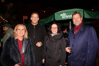 Four people smiling and holding drinks at an outdoor evening event, with umbrellas and festive lights in the background.