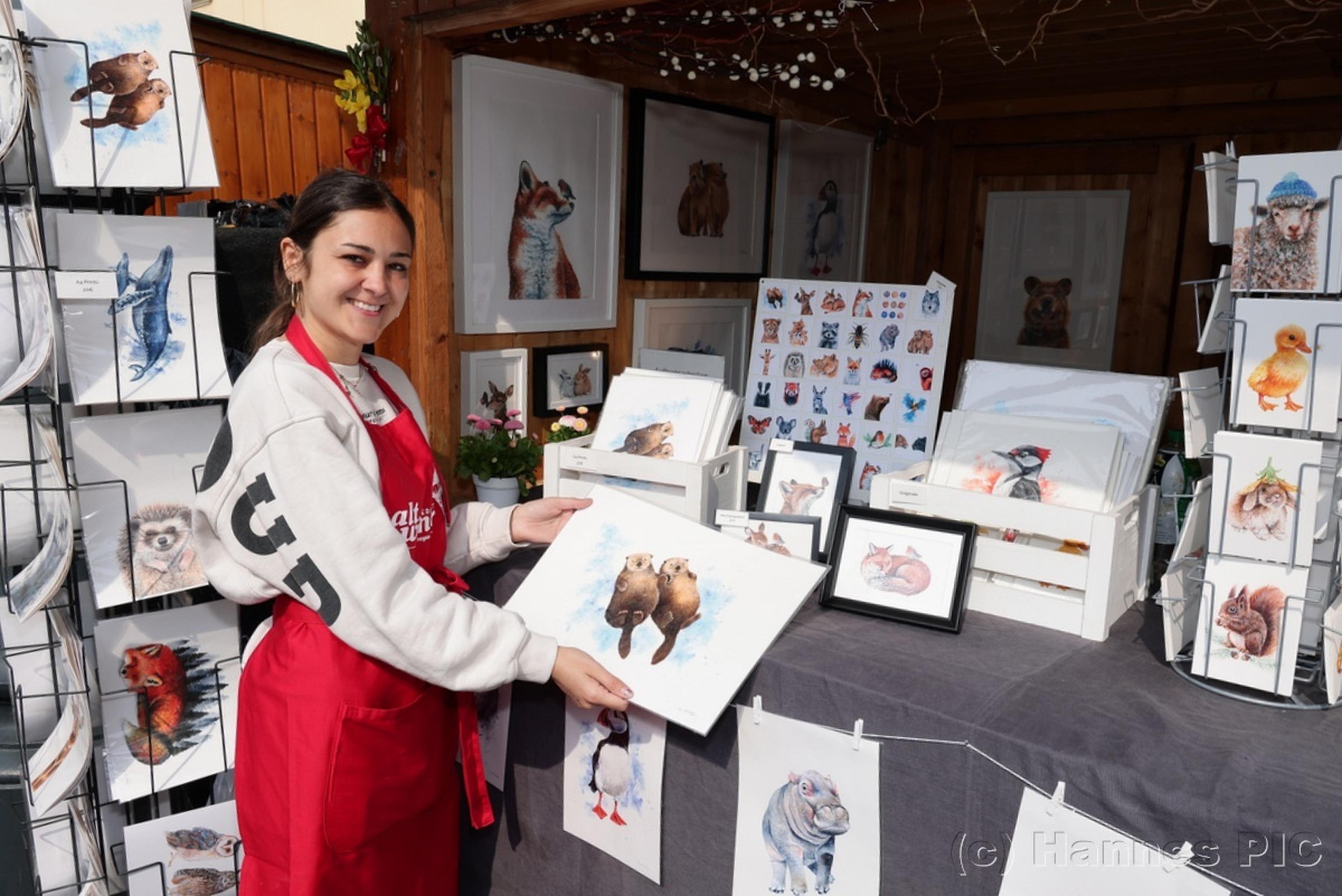 A smiling vendor in a red apron displays animal artwork at an outdoor market stall, featuring paintings of birds and other creatures.