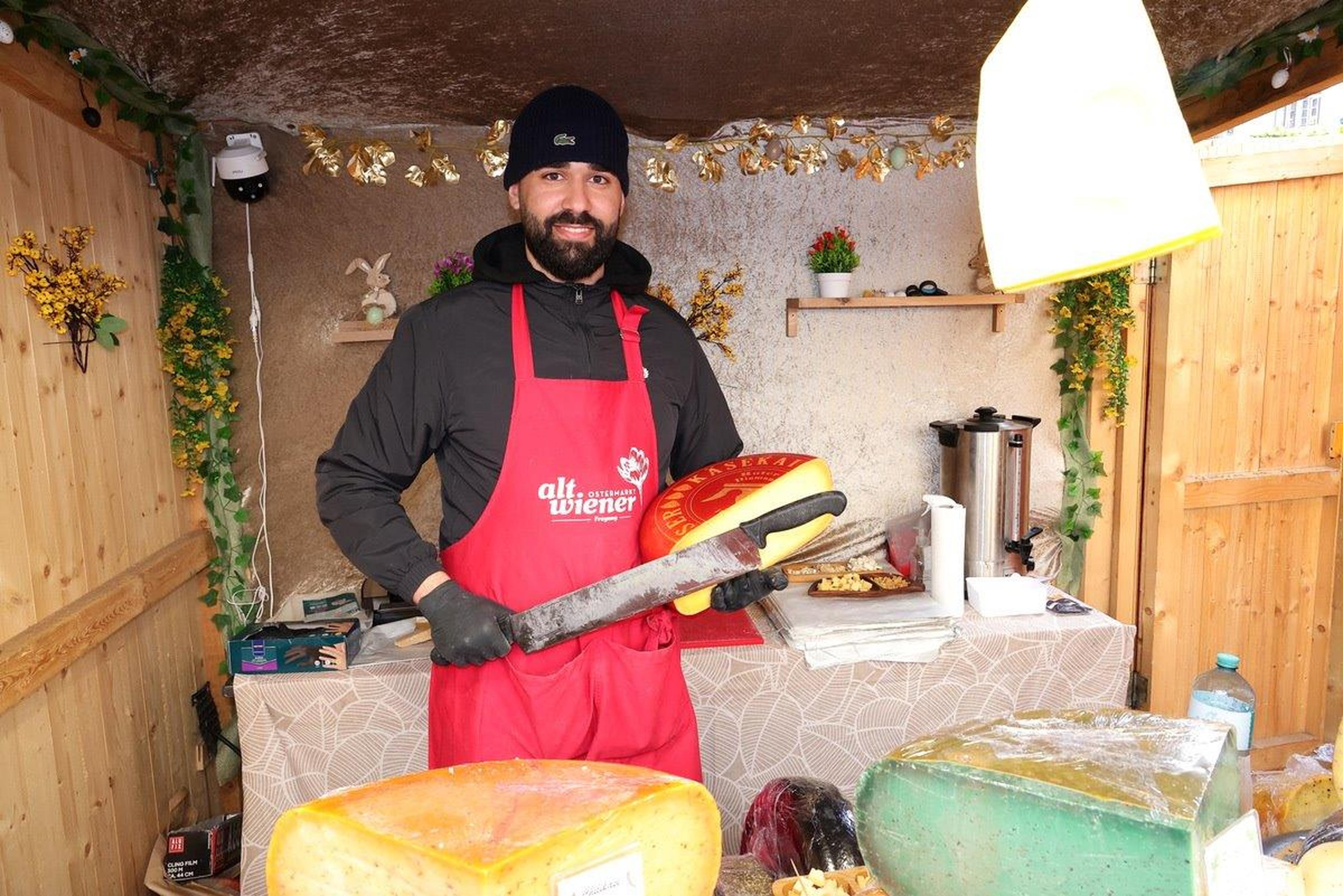 Man in a red apron and black jacket, holding a large knife and a wheel of cheese at an outdoor market stall with wooden walls and fairy lights.
