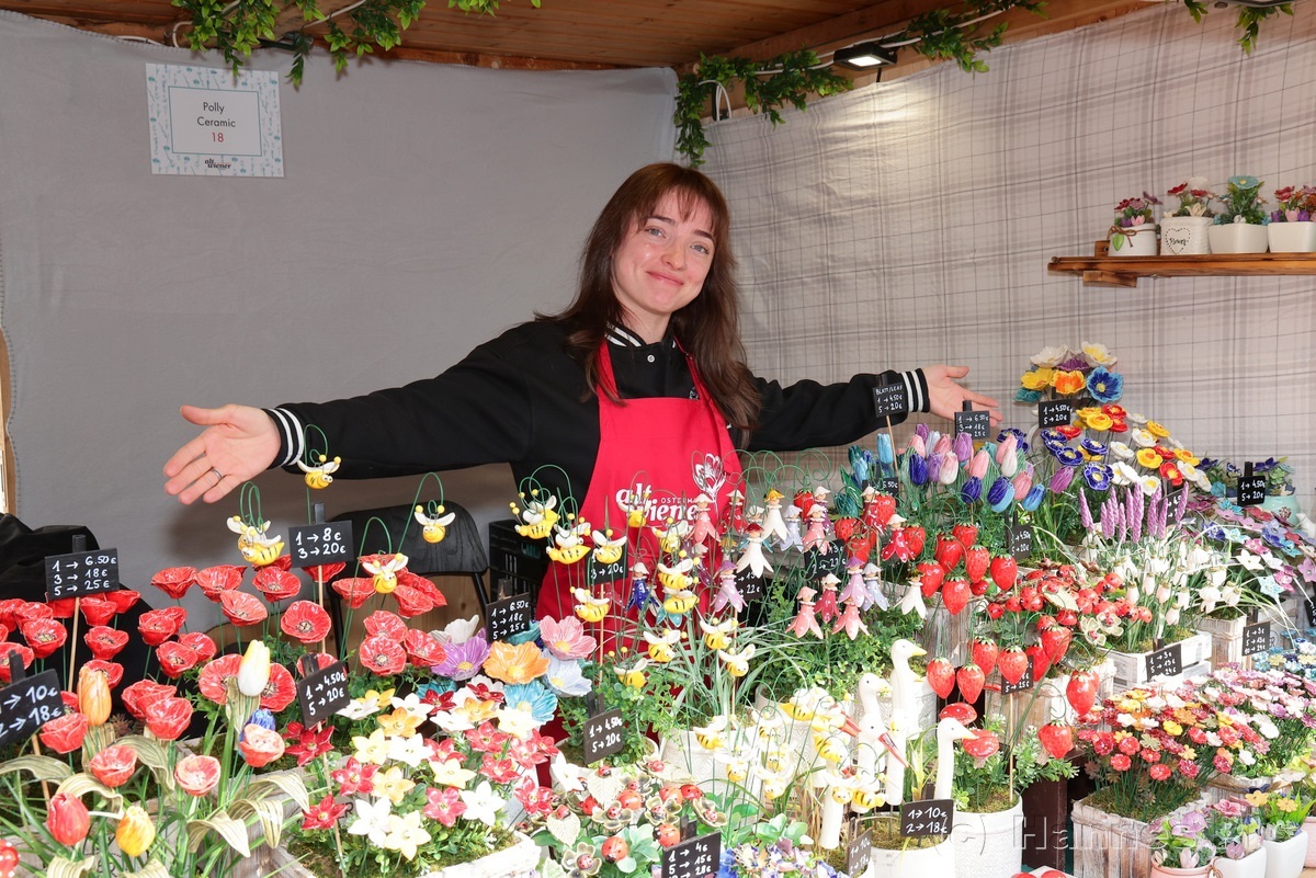 Smiling vendor in a red apron stands with arms open, showcasing a vibrant display of handcrafted, colorful clay flowers at a market stall.