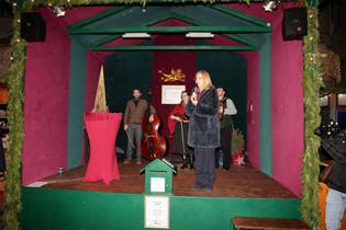 A woman speaks on a festive stage with musicians and a red draped table, surrounded by greenery and holiday decorations.