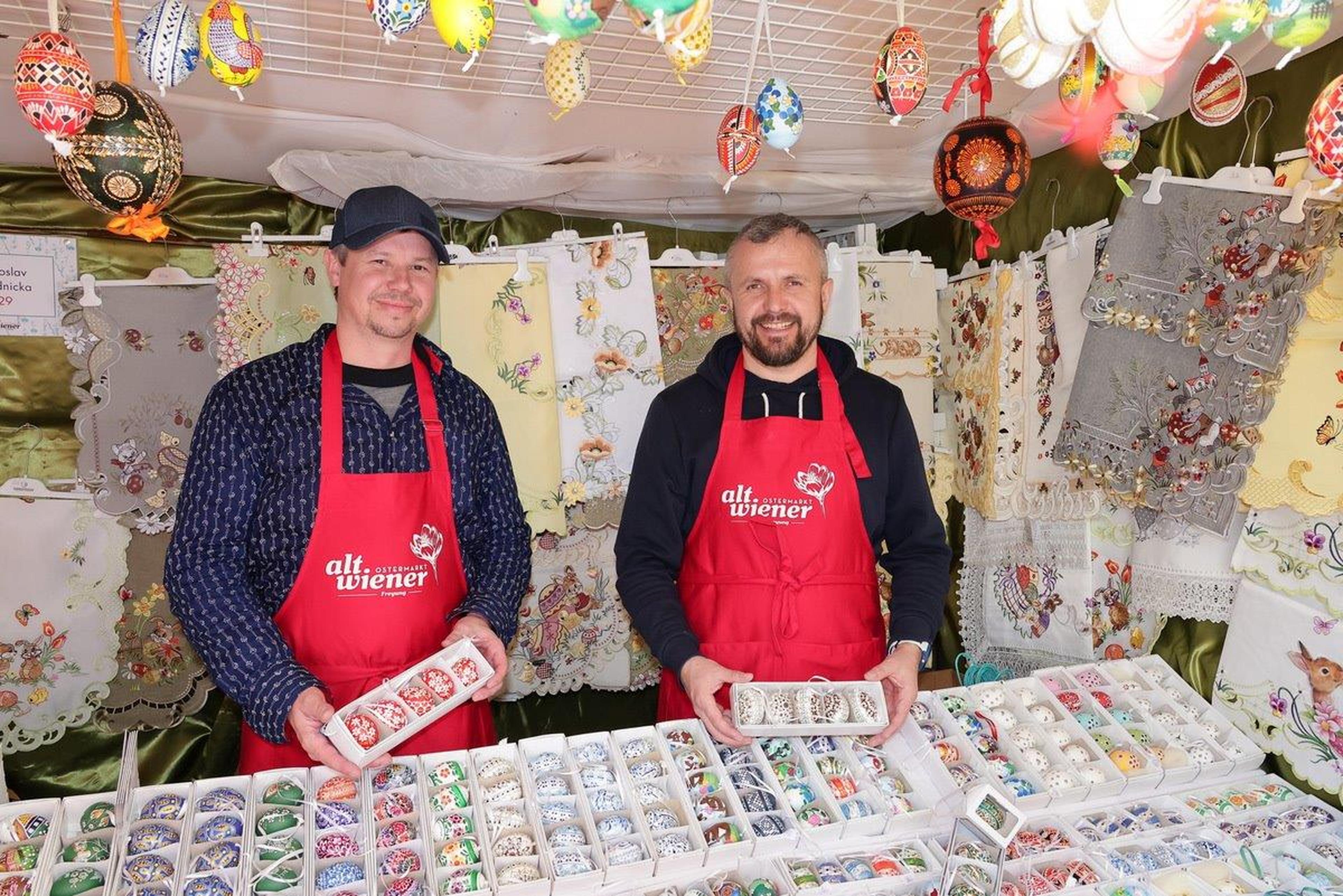 Two smiling vendors in red aprons holding decorated eggs at a festive market stall filled with colorful textiles and ornaments.
