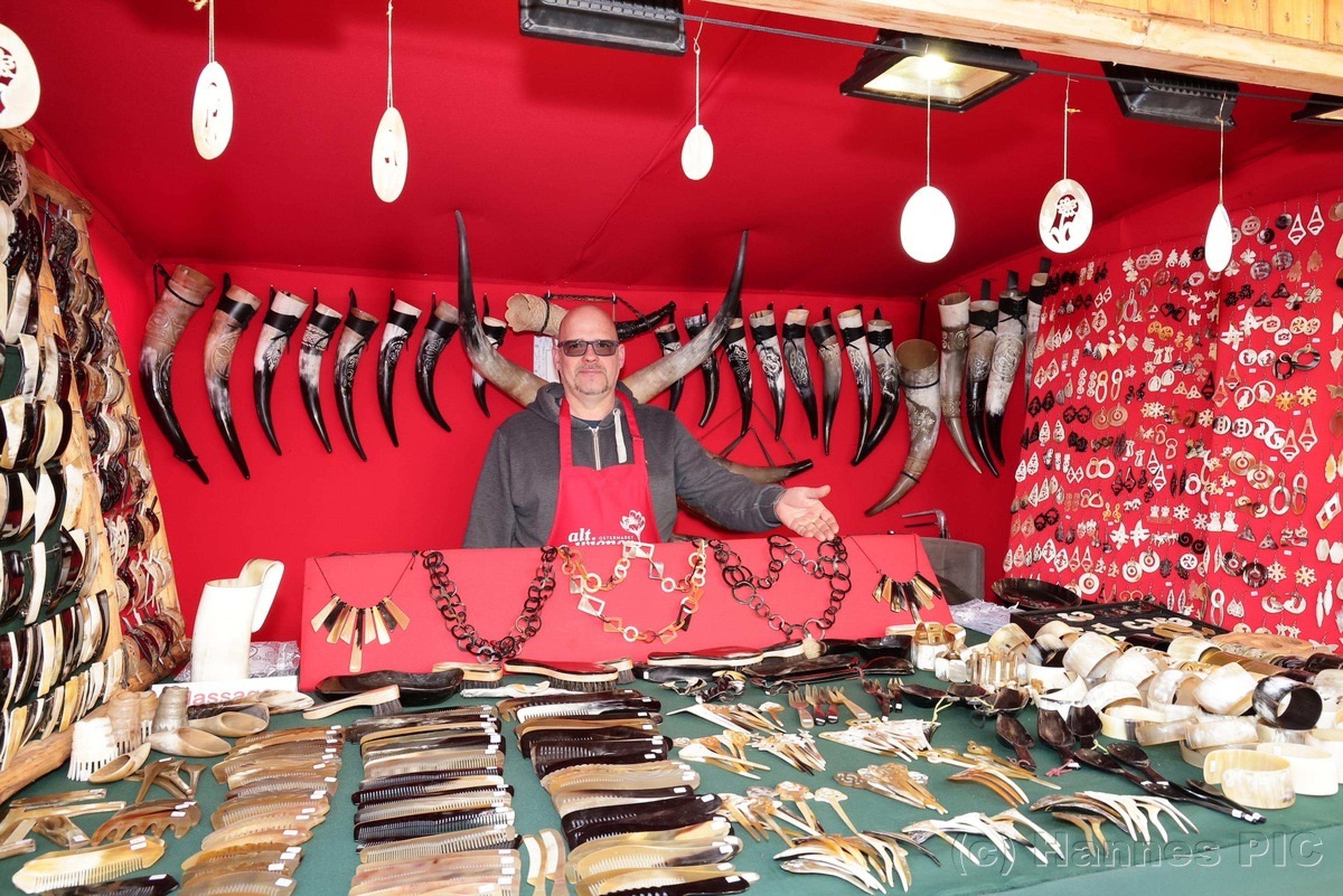 A vendor stands in a market stall displaying various horn crafts, necklaces, and combs against a red backdrop decorated with horns.