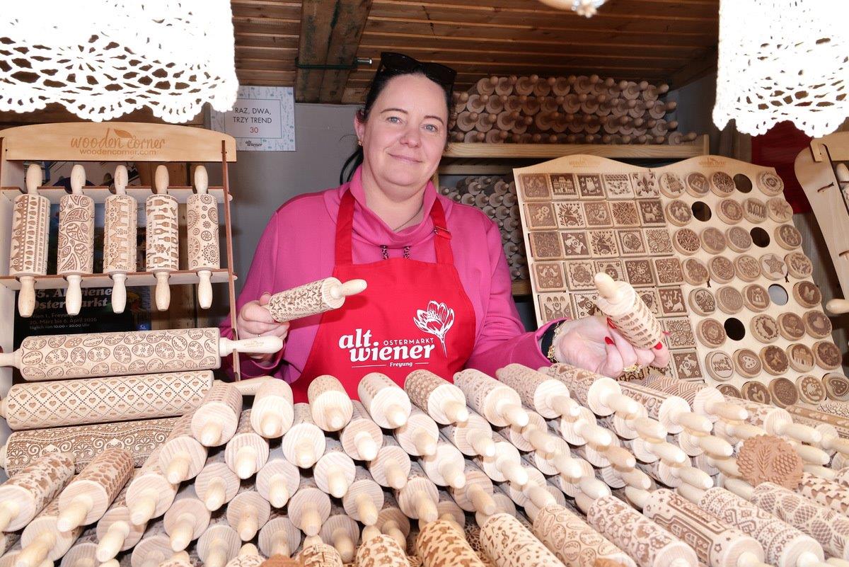 A woman in a red apron displays decorative rolling pins and engraved wooden items at a market stall, surrounded by intricately patterned designs.