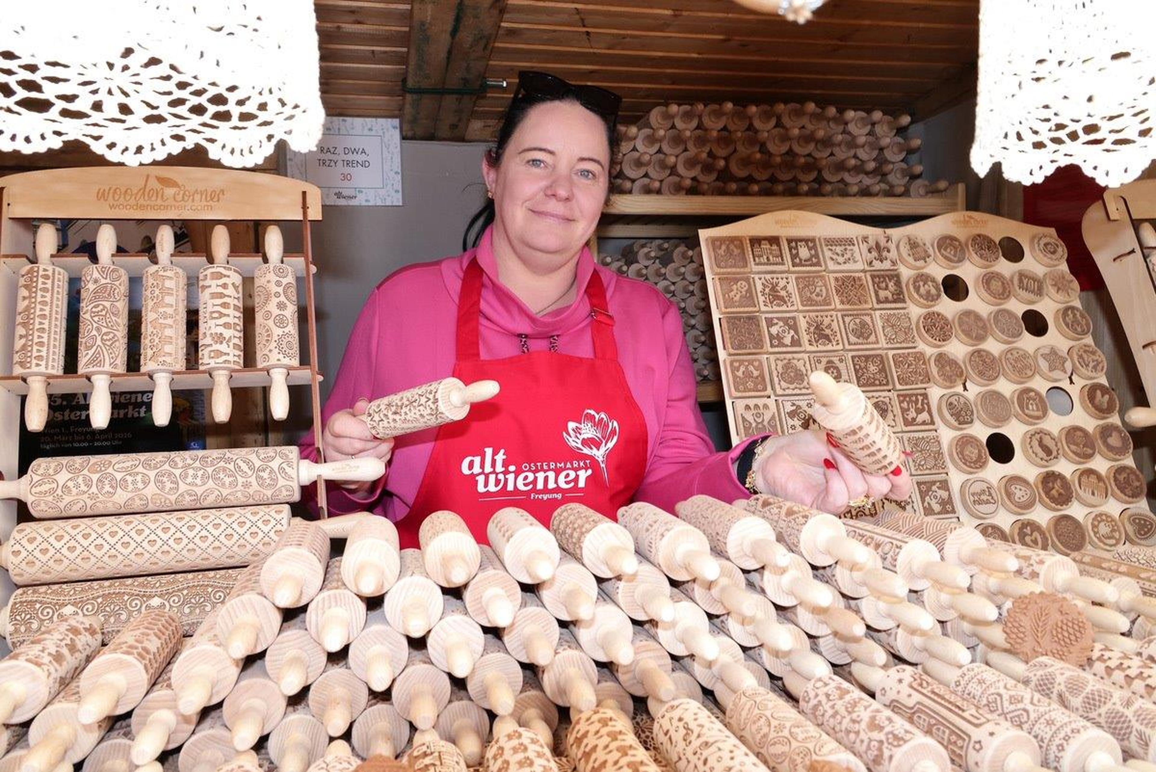 A woman in a red apron displays decorative rolling pins and engraved wooden items at a market stall, surrounded by intricately patterned designs.