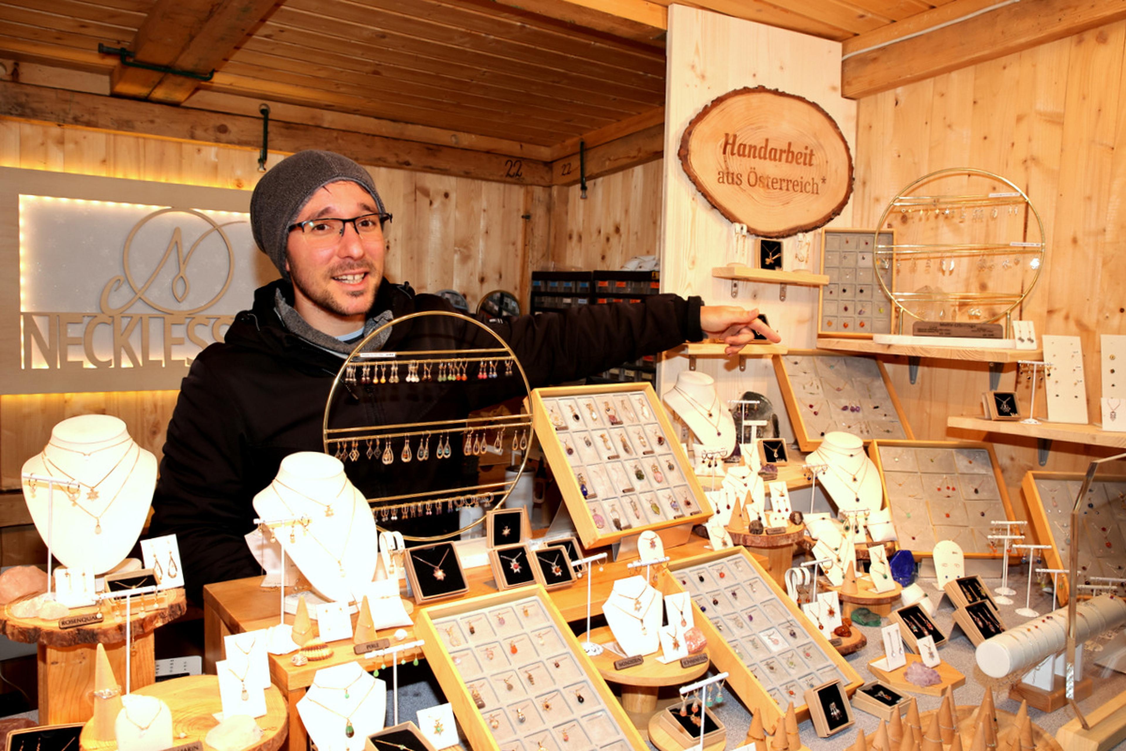 A vendor in a cozy wooden booth displays handcrafted jewelry, with necklaces and rings. A sign reads "Handarbeit aus Österreich" (Handmade from Austria).
