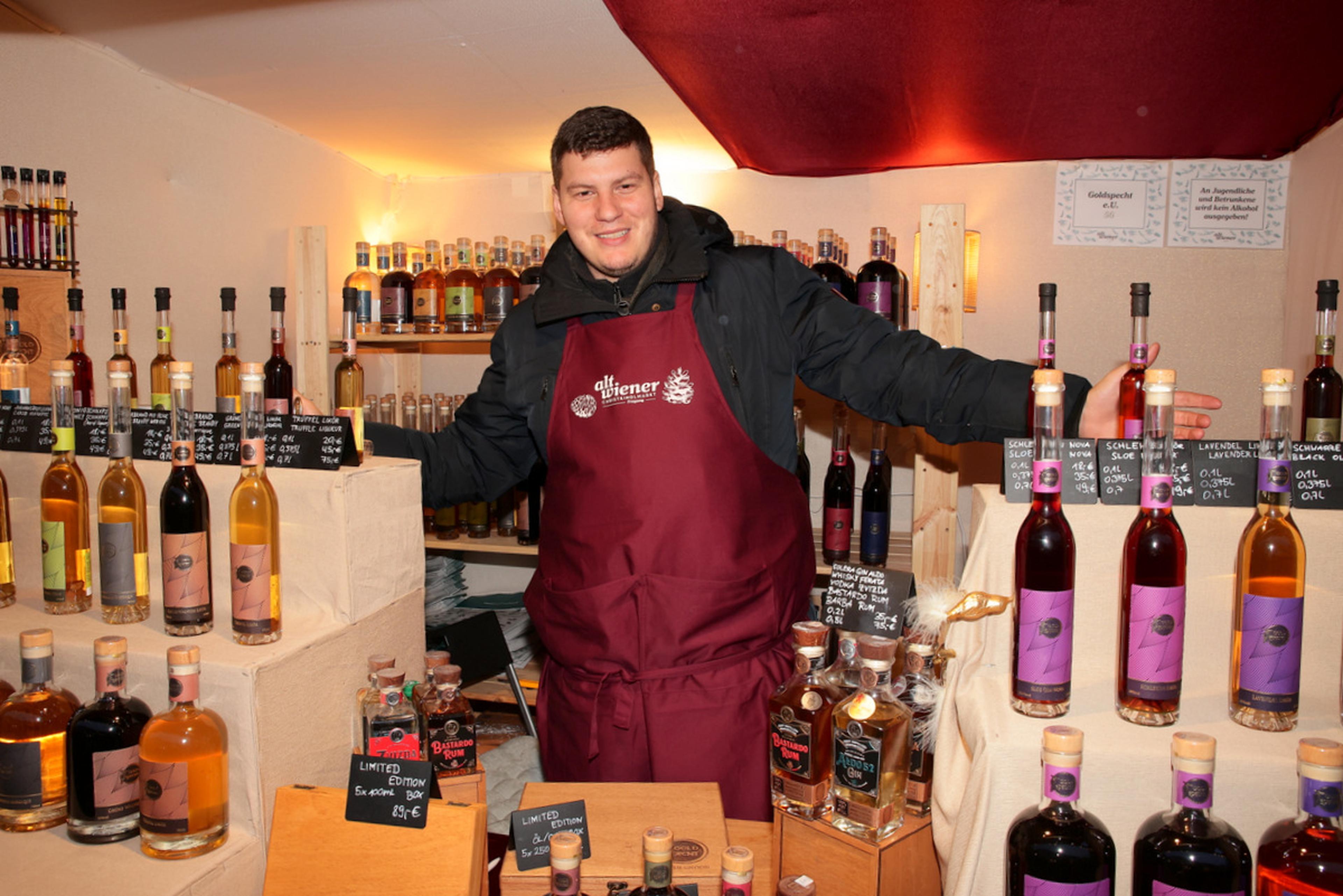 Man in a maroon apron stands smiling behind a display of various bottled beverages at a market stall.