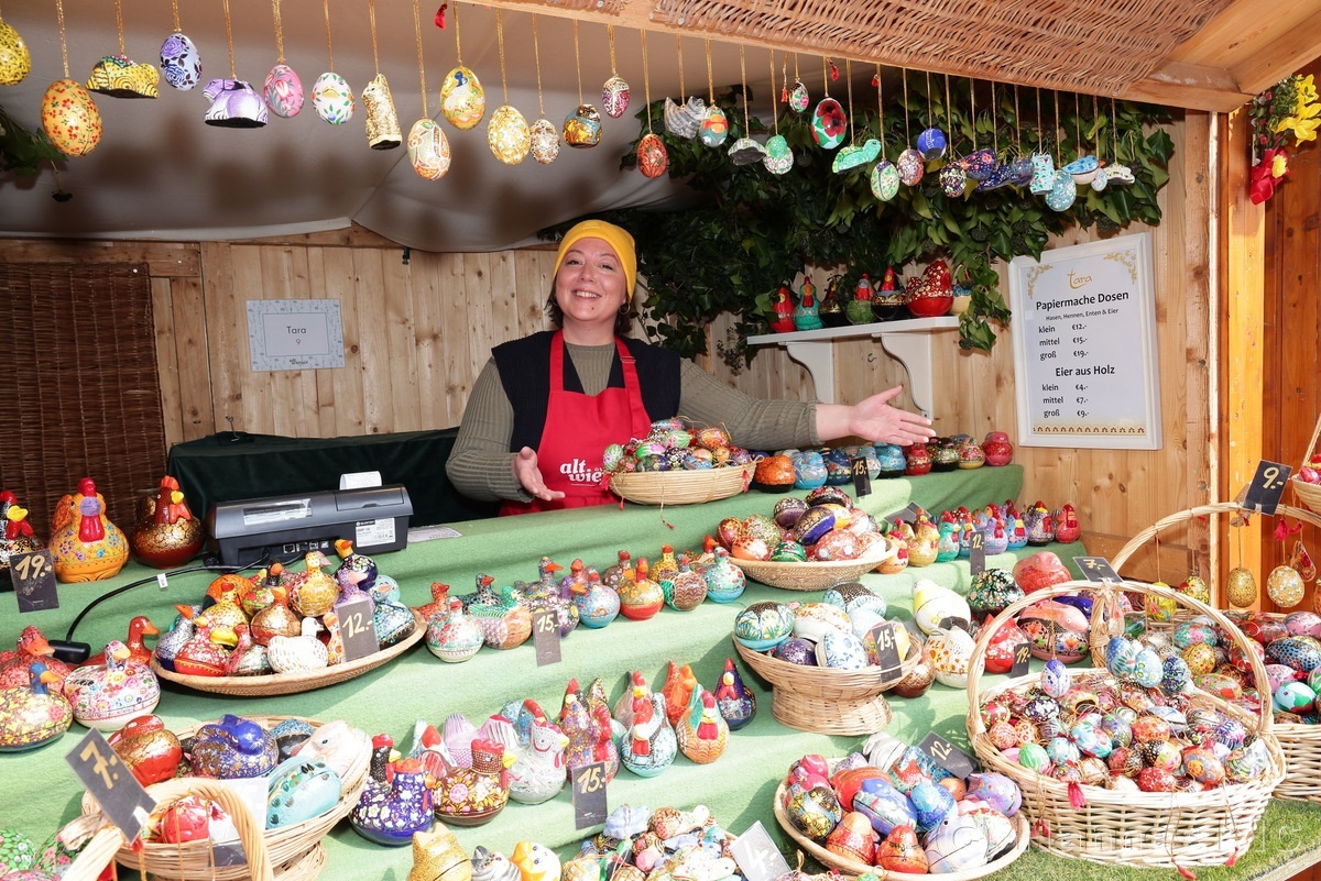 Seller in a festive booth smiling, showcasing colorful, decorative eggs and ornaments on display, under a wooden roof at a market stall.