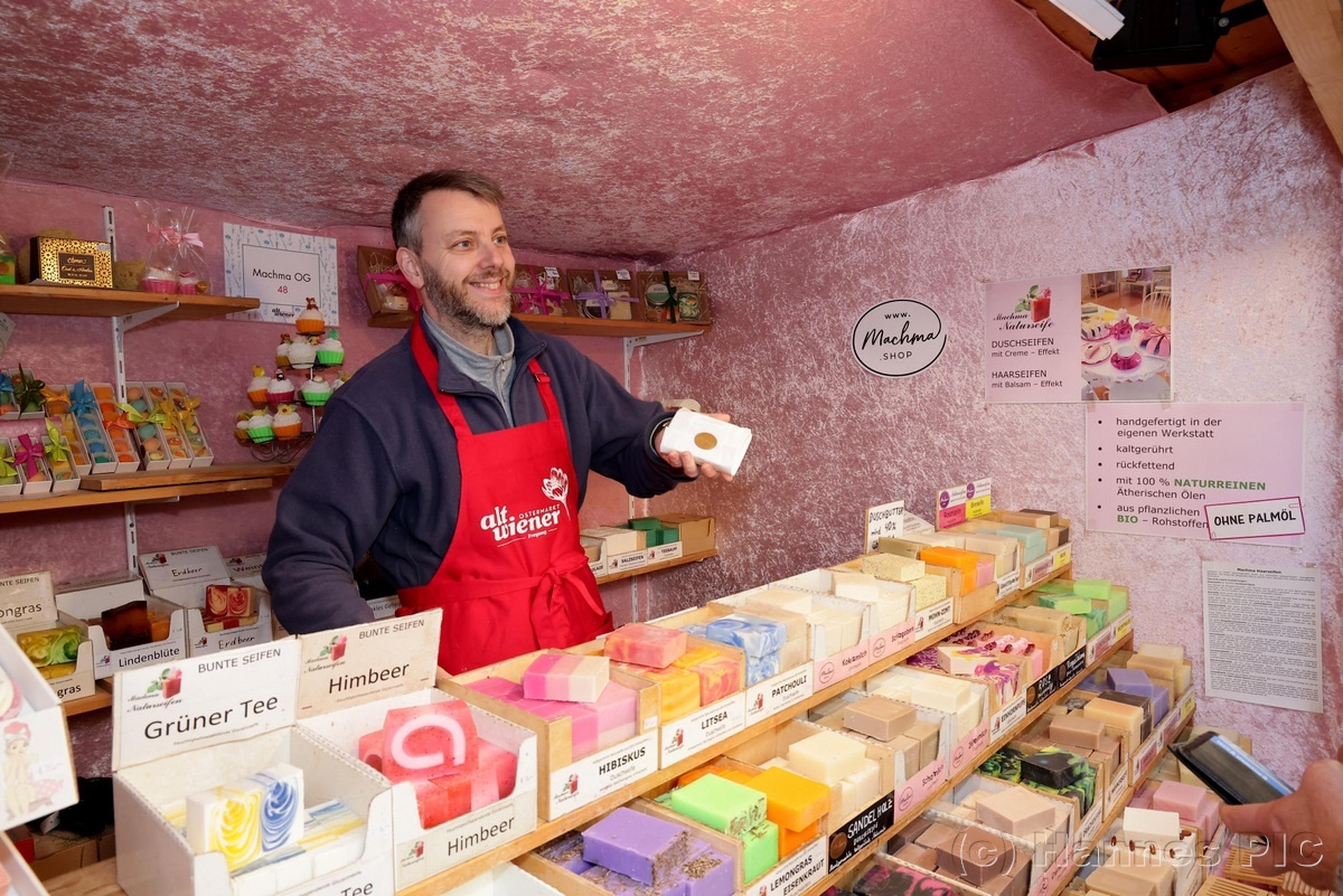 Smiling vendor in a red apron displays colorful handmade soap bars in a cozy market stall with pink walls.