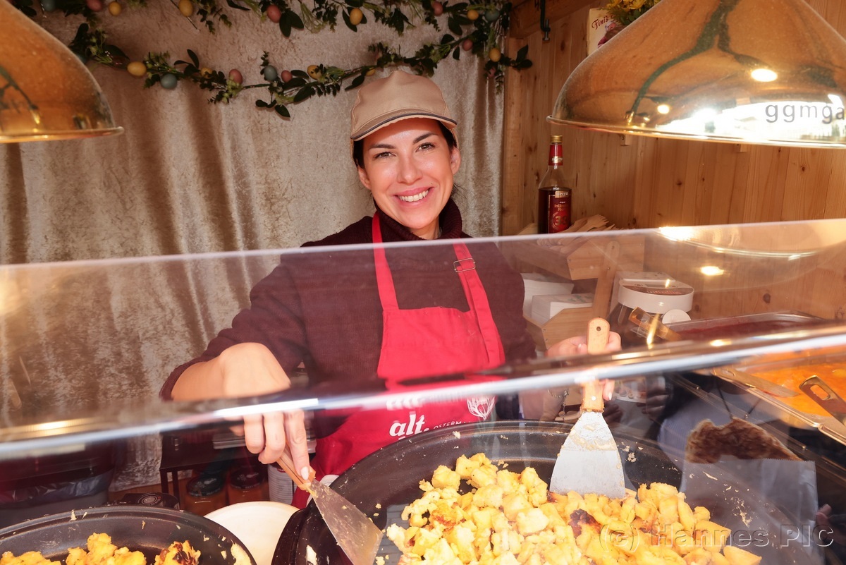 Smiling vendor in a red apron serves fried potatoes at a food stall, surrounded by decorative elements and cooking utensils.