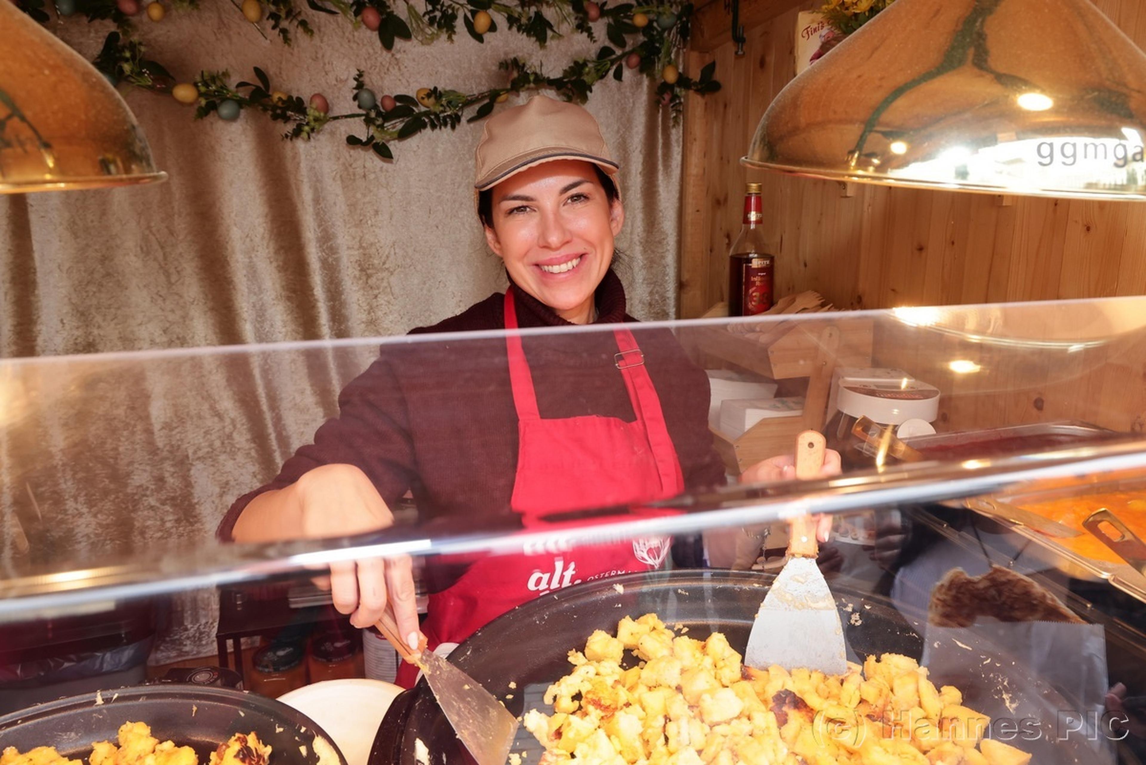 Smiling vendor in a red apron serves fried potatoes at a food stall, surrounded by decorative elements and cooking utensils.