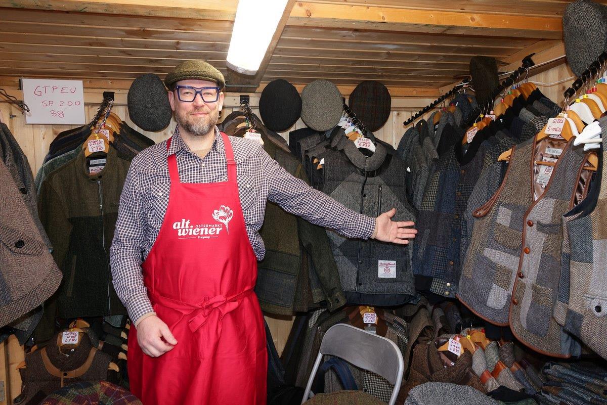 Man in a red apron and cap displaying various tweed jackets and vests in a wooden booth.