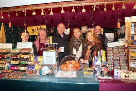 A group of six people stand smiling behind a festive market stall. Shelves display colorful chocolates and sweets. The setting is warm and inviting, adorned with twinkling lights and small decorations. A glass case showcases fresh rolls. A sign indicates accepting donations. The atmosphere is cheerful and welcoming.