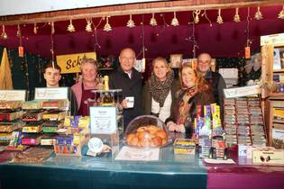 A group of six people stand smiling behind a festive market stall. Shelves display colorful chocolates and sweets. The setting is warm and inviting, adorned with twinkling lights and small decorations. A glass case showcases fresh rolls. A sign indicates accepting donations. The atmosphere is cheerful and welcoming.